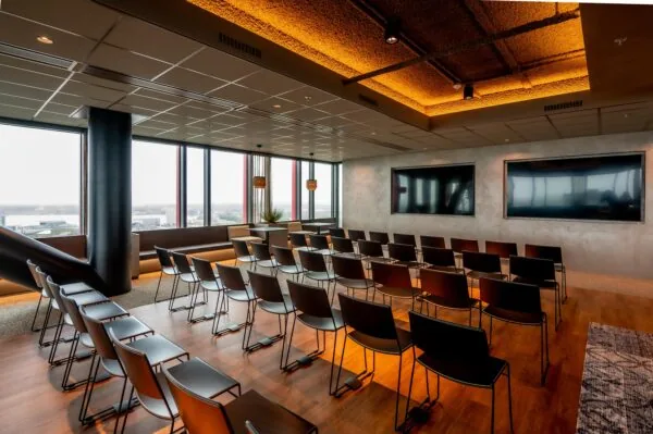 Modern conference room with rows of black chairs, two large screens, and panoramic city views through tall windows.