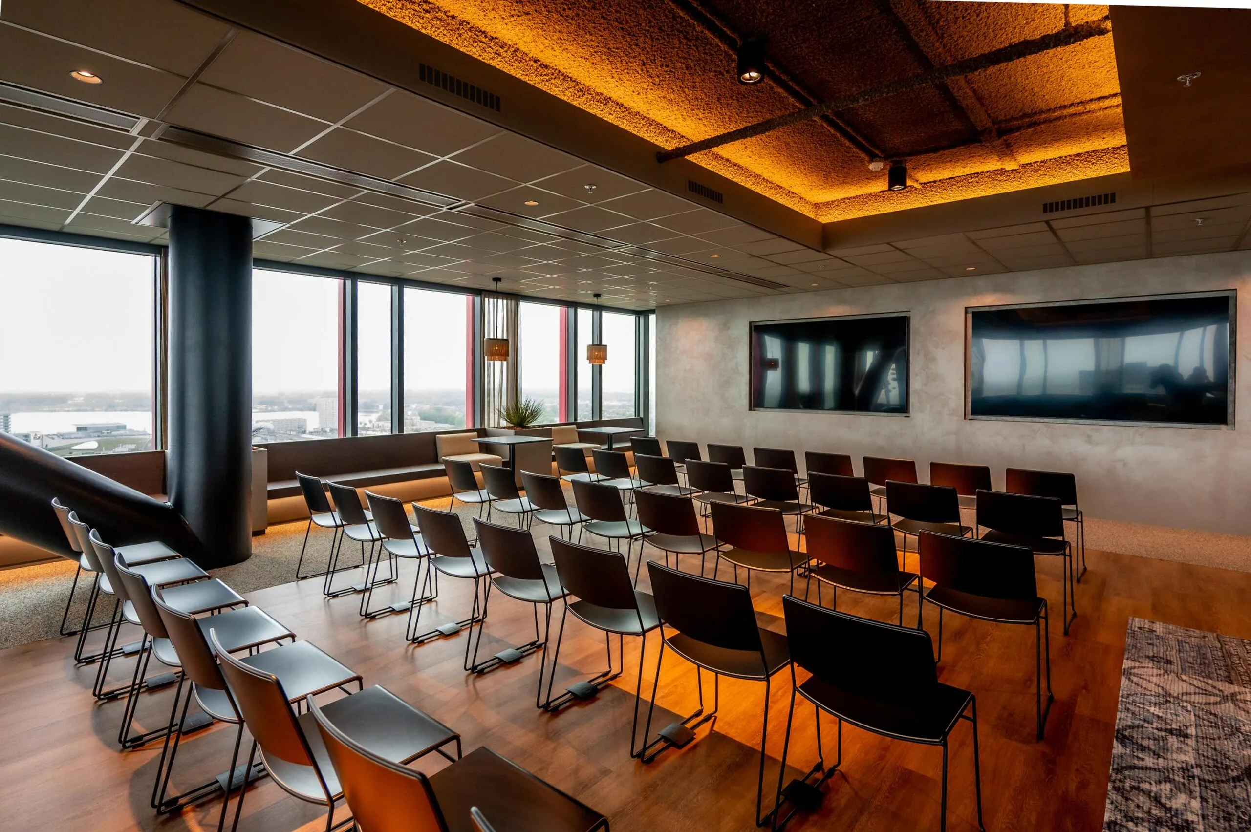 Modern conference room with rows of black chairs, two large screens, and panoramic city views through tall windows.