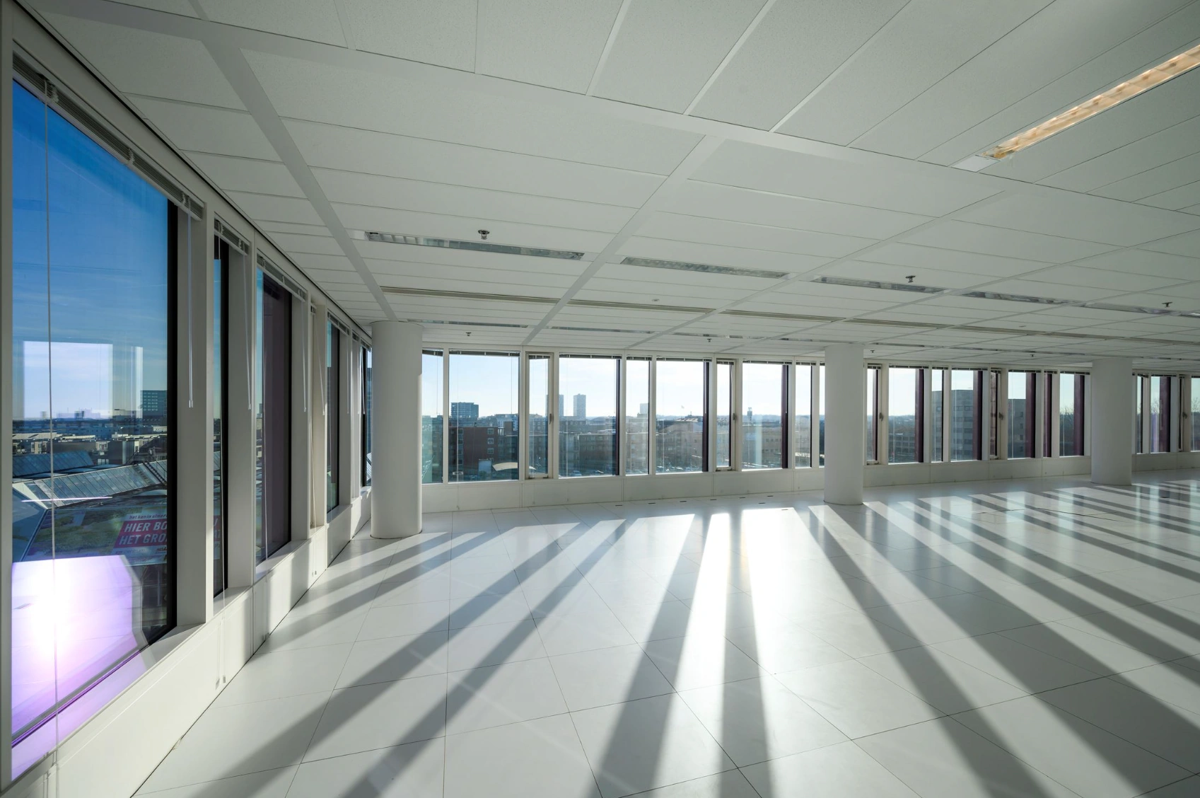 Bright, empty office space on P.J. Oudweg with large windows offering a panoramic city view and casting long shadows on the floor.