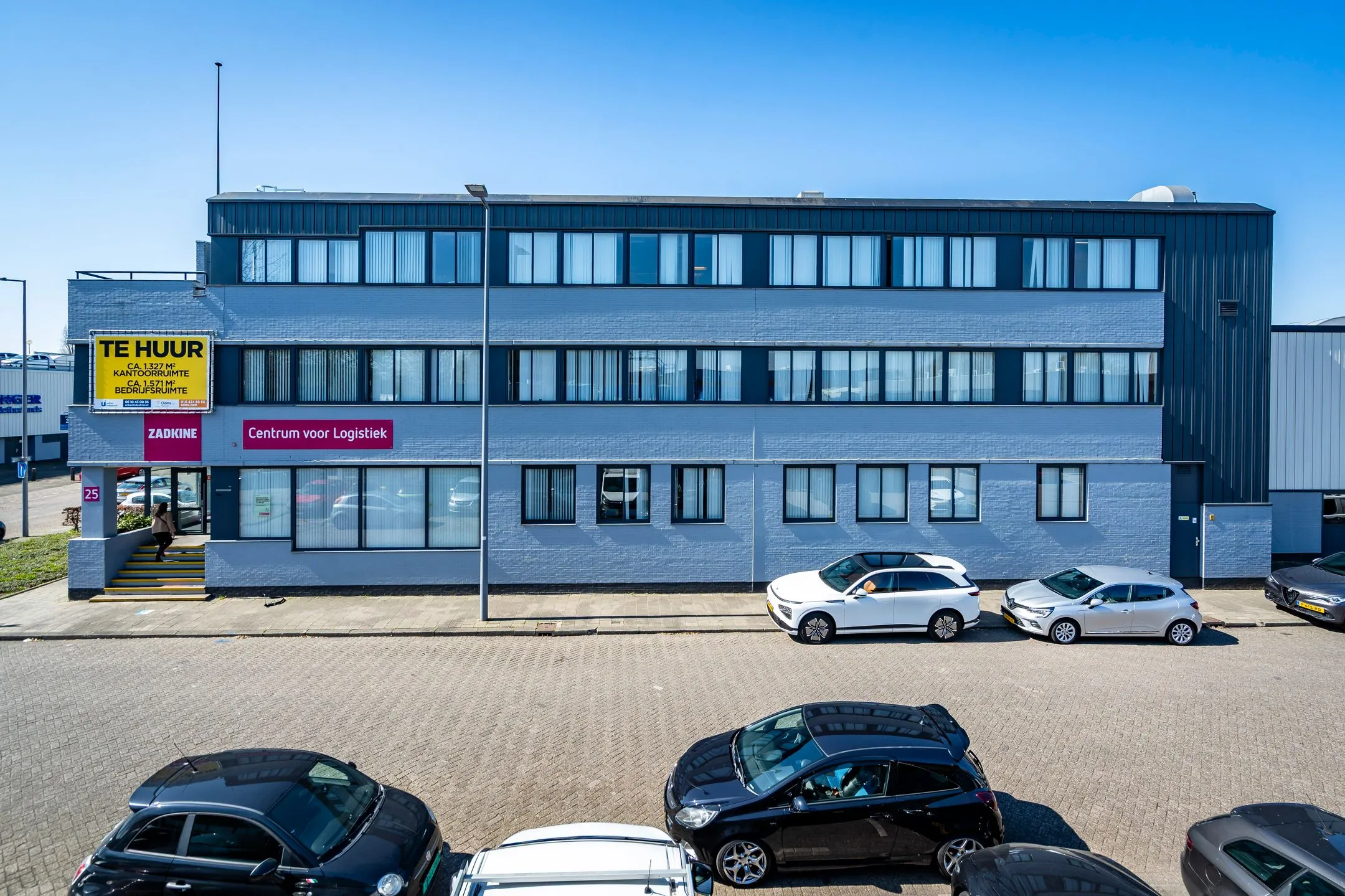 Modern three-story office and logistics building on Aluminiumstraat with cars parked in front and a "For Rent" sign displayed.
