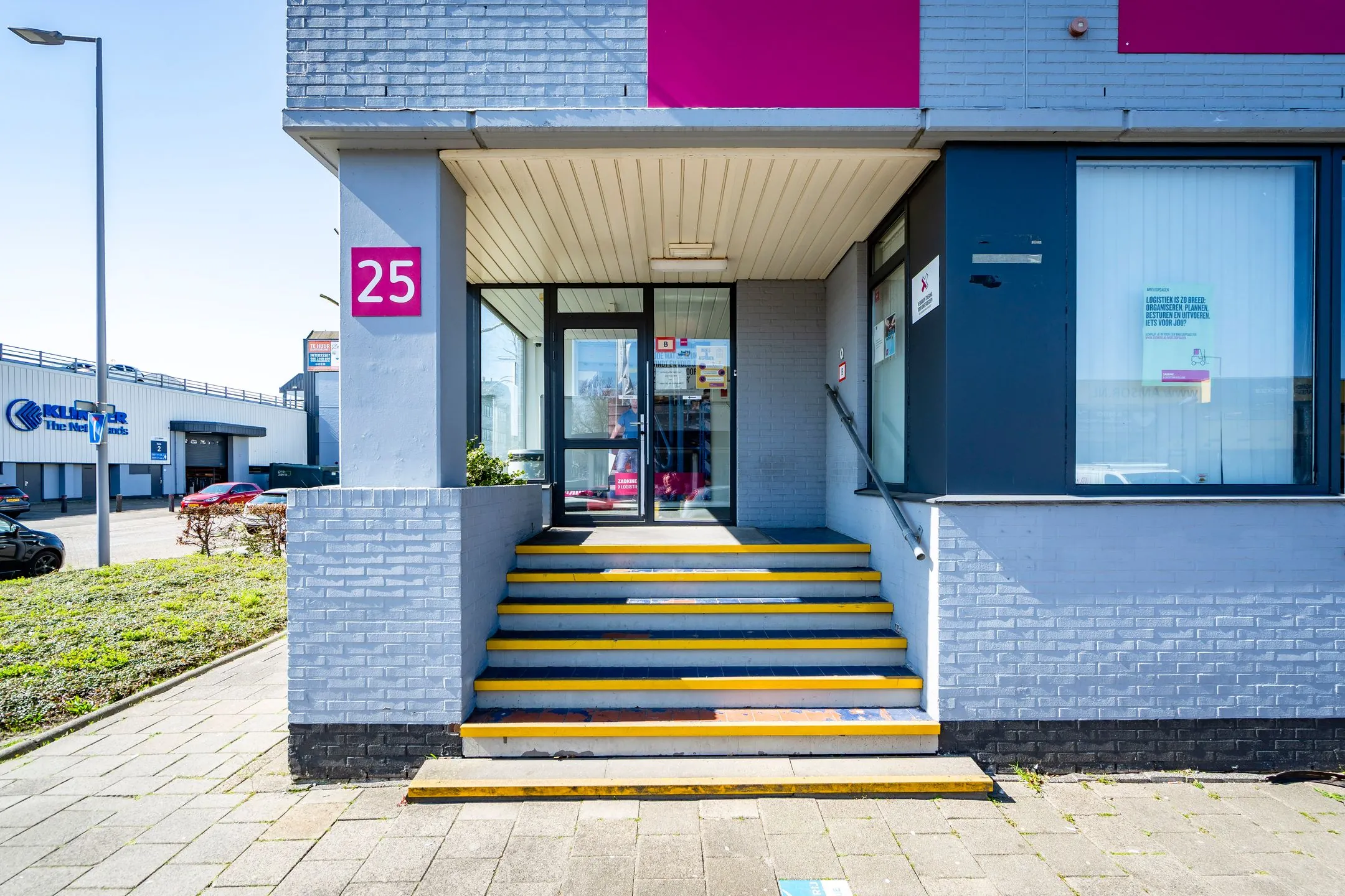 Entrance of a gray commercial building at Aluminiumstraat 25 with yellow-striped steps and glass doors.