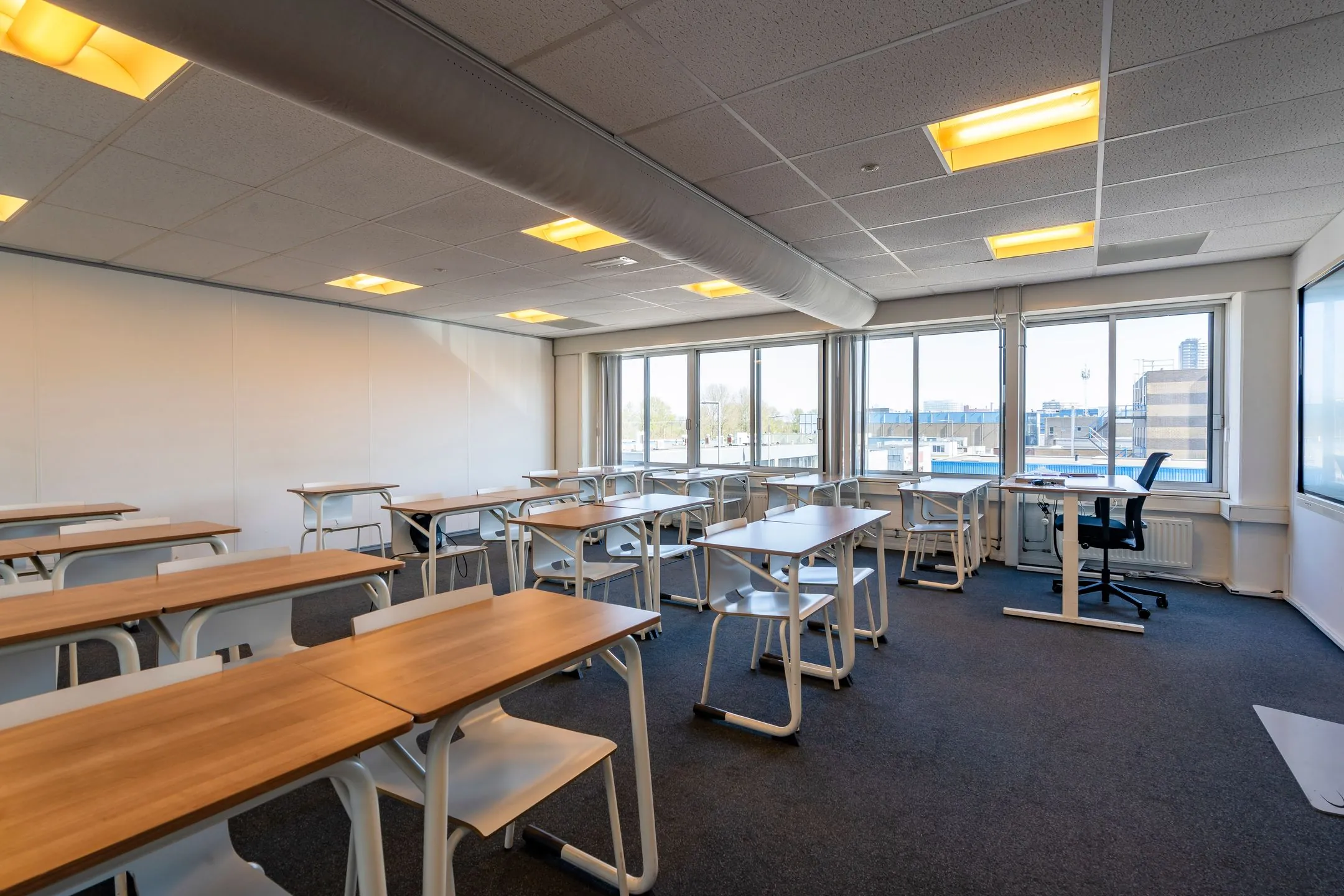 Modern classroom with wooden desks, white chairs, and large windows overlooking an urban area on Aluminiumstraat.