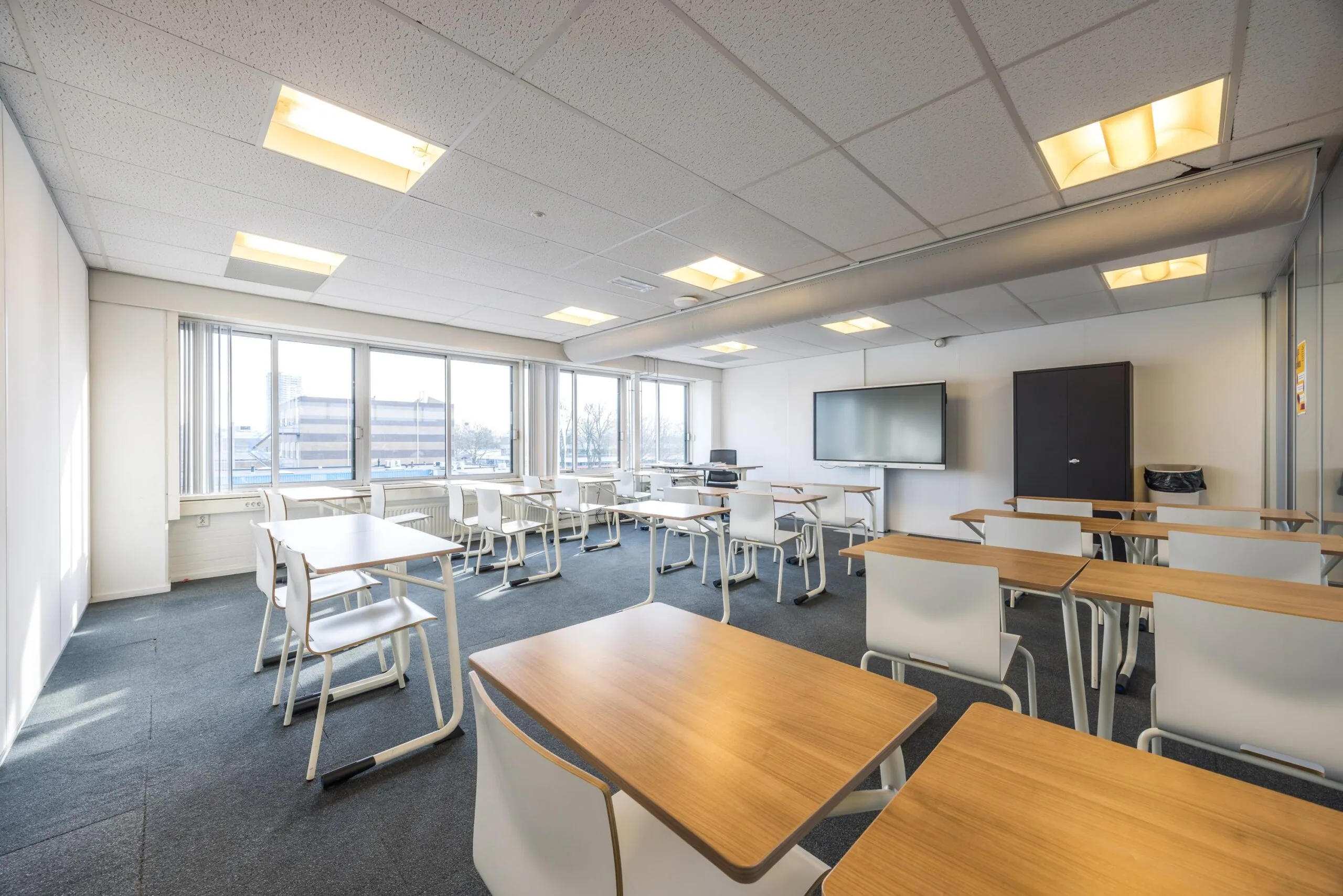 Modern classroom at Aluminiumstraat with wooden desks, white chairs, large windows, and a digital screen.