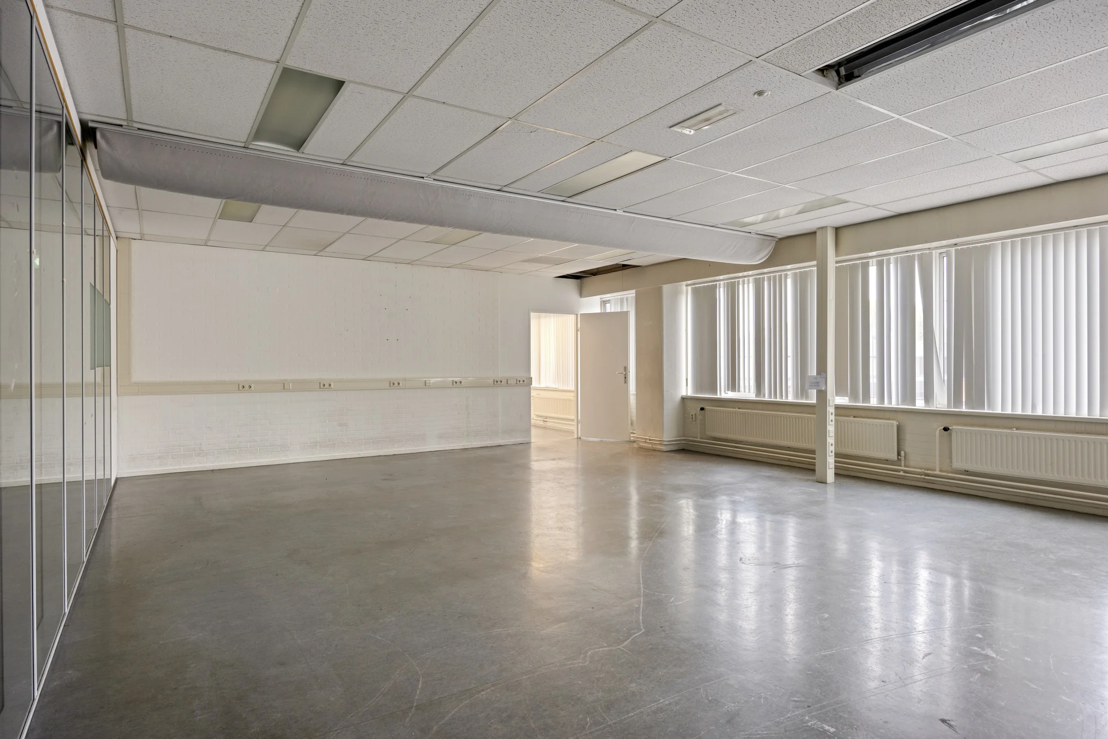 Empty commercial space on Aluminiumstraat with large windows, fluorescent ceiling lights, and polished concrete flooring.