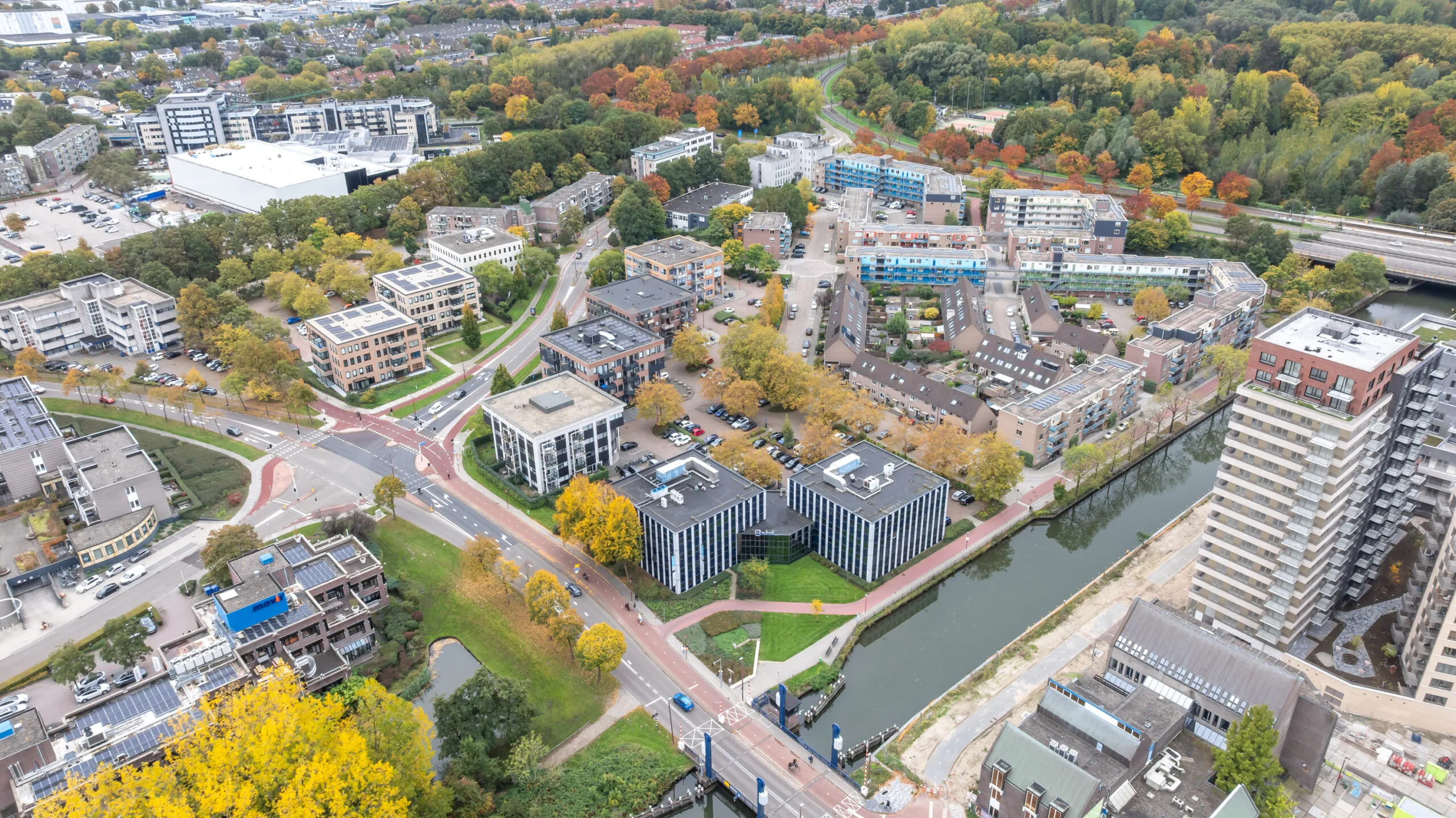 Aerial view of the Krijtwal area featuring office buildings, residential apartments, roads, and a canal surrounded by autumn-colored trees.