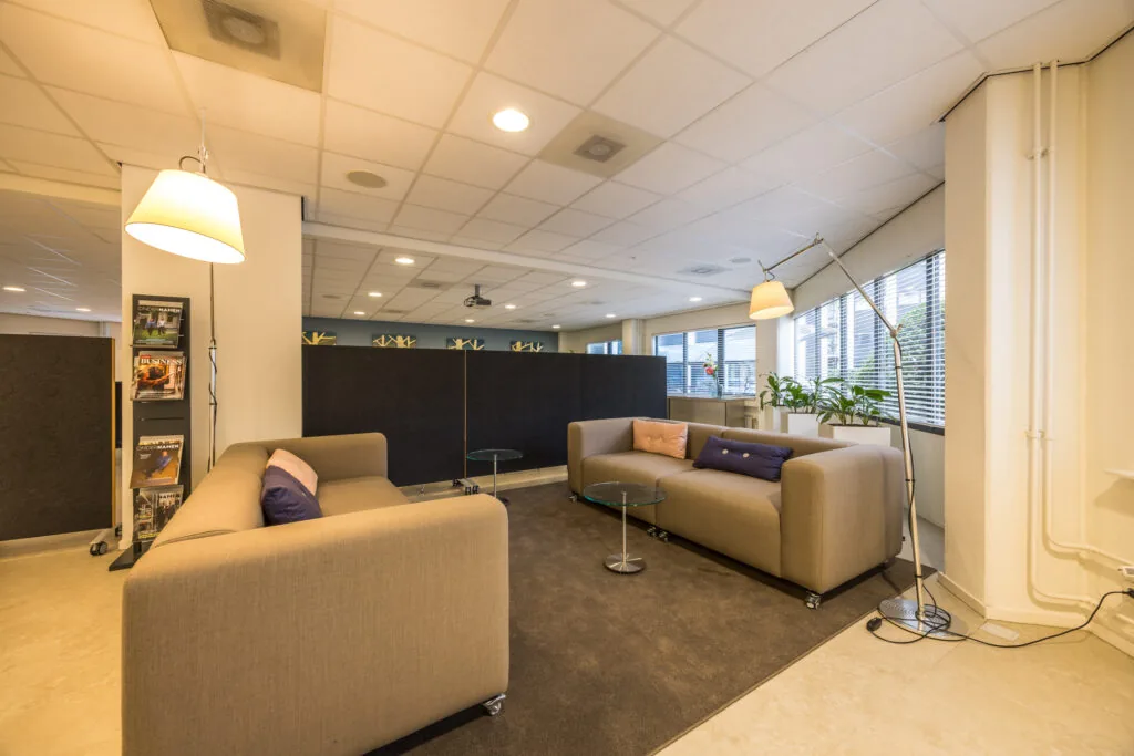 Modern office lounge with beige sofas, glass coffee tables, and large windows providing natural light.