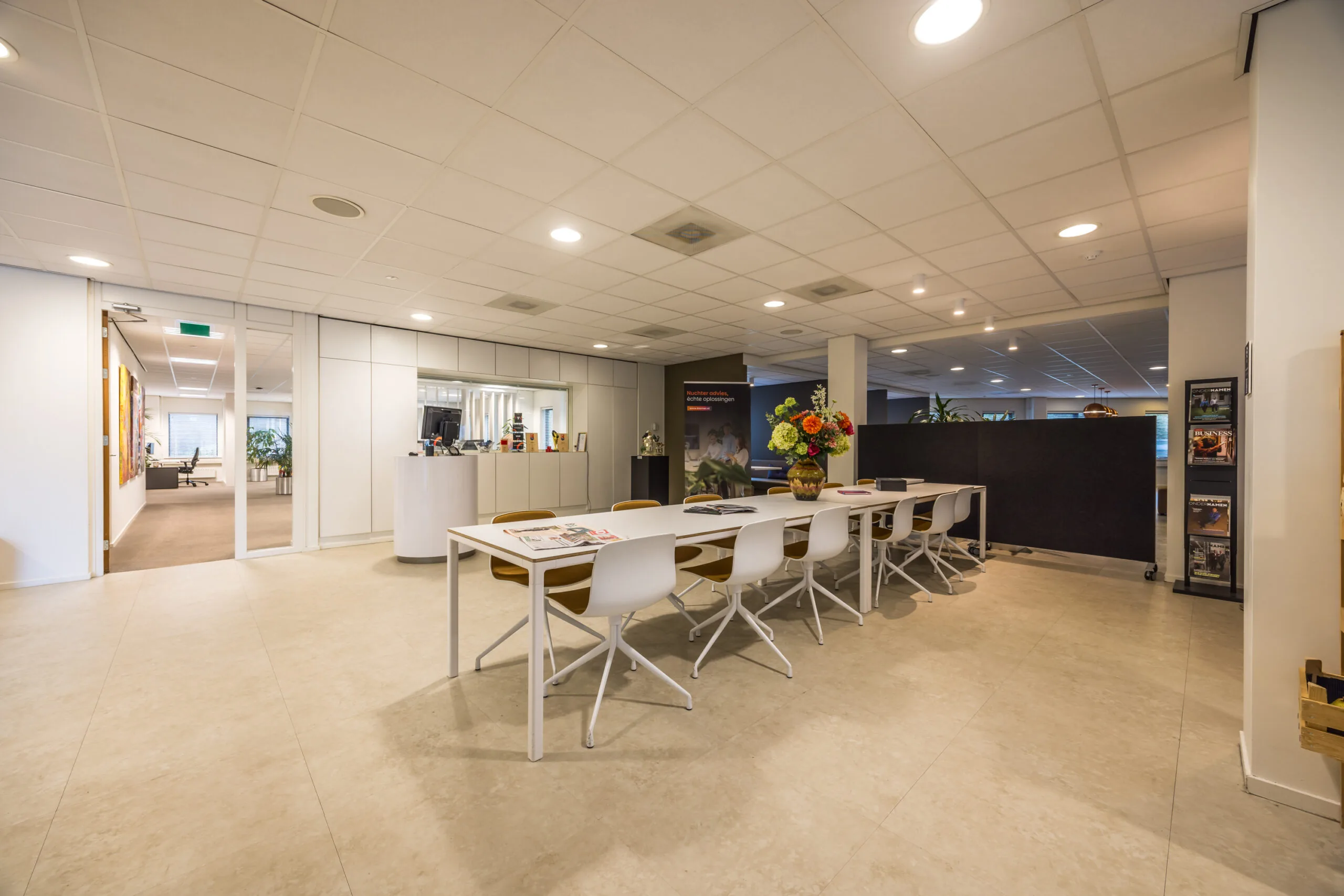 Modern office space with a long white table, white chairs, and a vase of colorful flowers in the center.