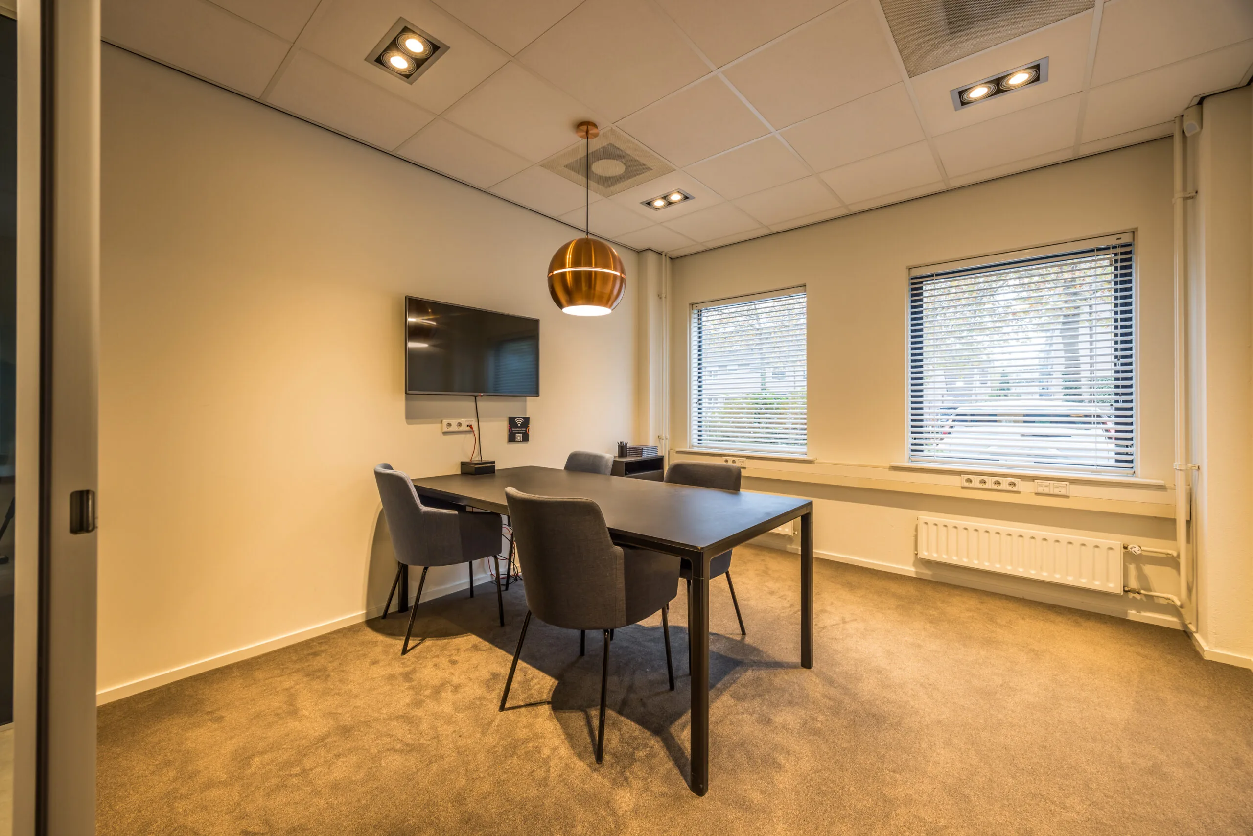 Modern meeting room at Krijtwal with a black table, four grey chairs, a wall-mounted TV, and large windows with blinds.