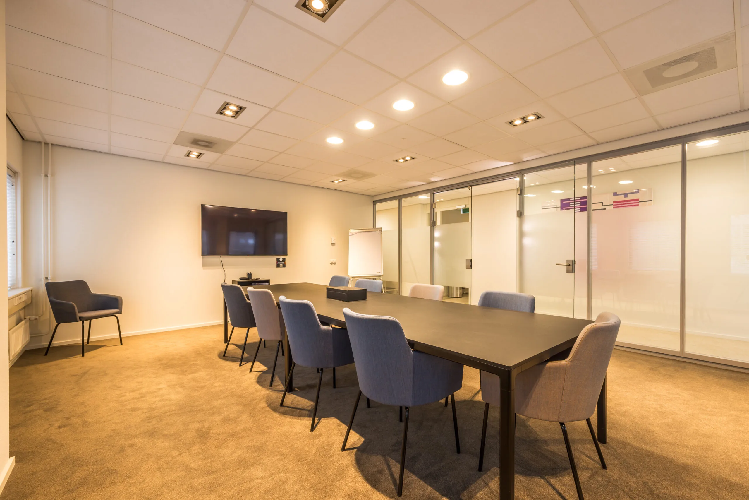Modern conference room at Krijtwal featuring a long table, upholstered chairs, a wall-mounted screen, and glass partition walls.