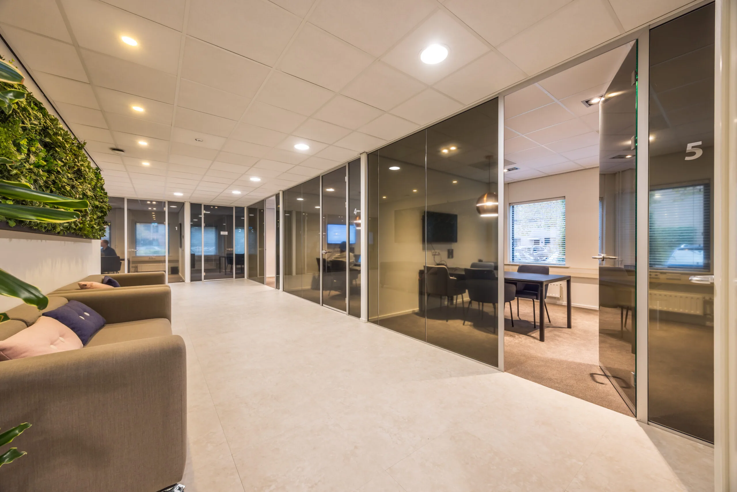 Modern office hallway with glass-walled meeting rooms and a seating area featuring a green plant wall.