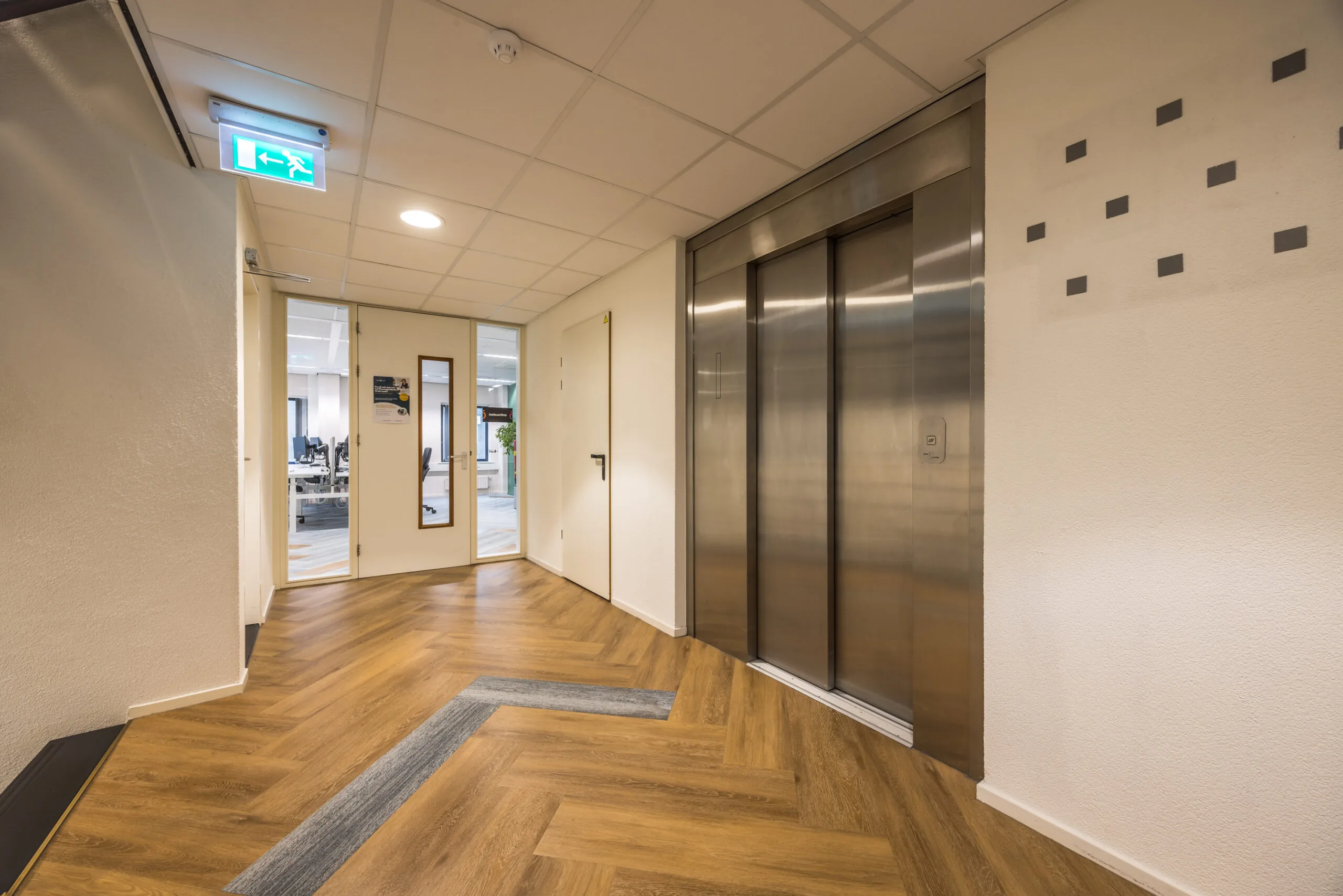Modern office hallway with wooden herringbone flooring, a stainless steel elevator, and a glass door leading to a workspace in Krijtwal 176.