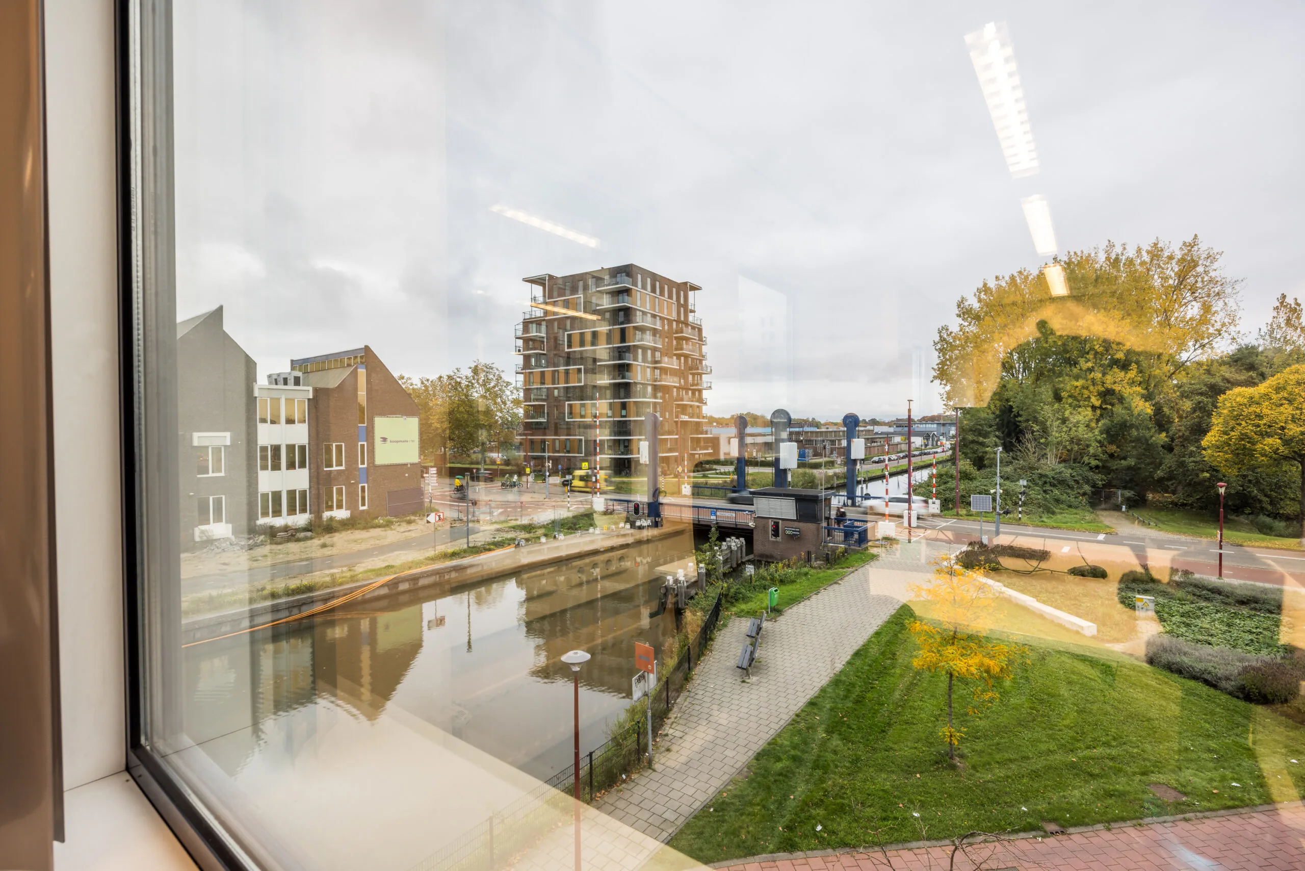 View through a window showing the Krijtwal canal, a modern apartment building, and a bridge in an urban setting with greenery.