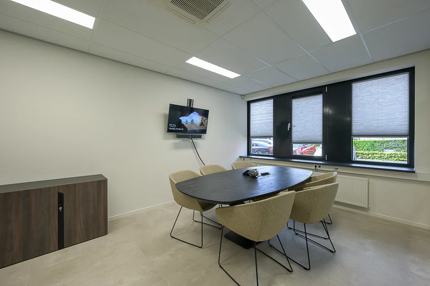 Modern meeting room at Ebweg with a black oval table, six beige chairs, a wall-mounted screen, and large windows with blinds.