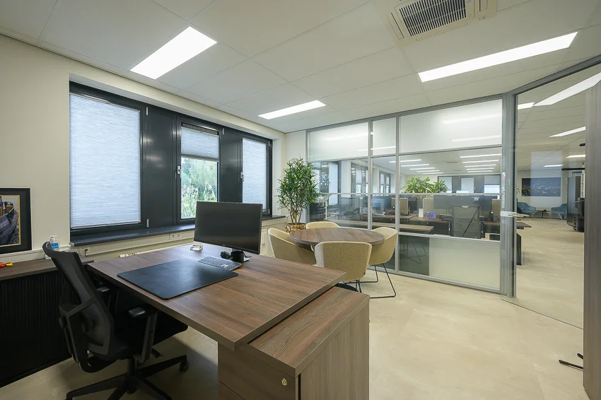 Modern office space with a wooden desk, ergonomic chair, lounge chairs, and glass walls overlooking an open-plan workspace.