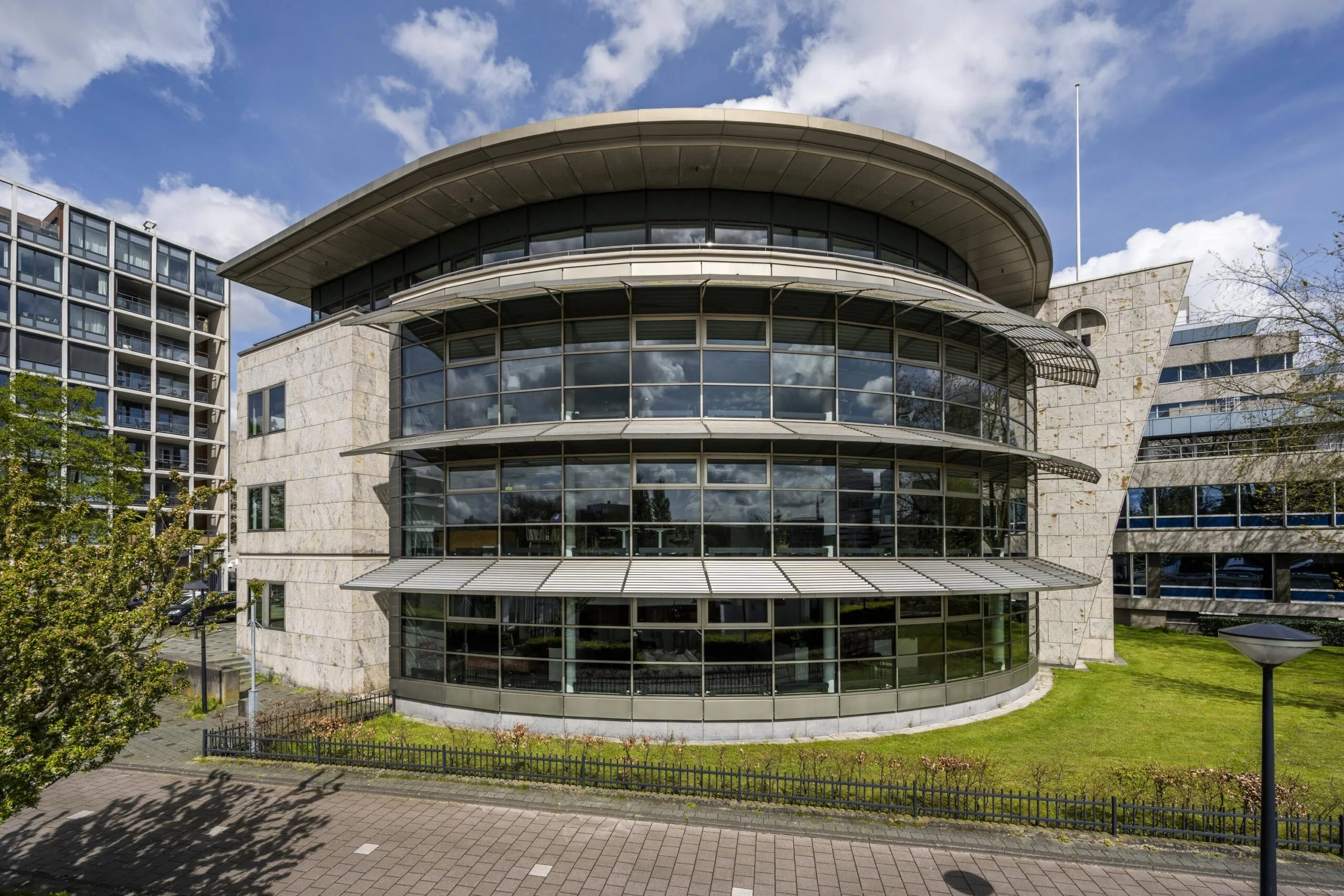 Modern curved office building "De Omval" with large glass windows and a stone facade under a partly cloudy sky.