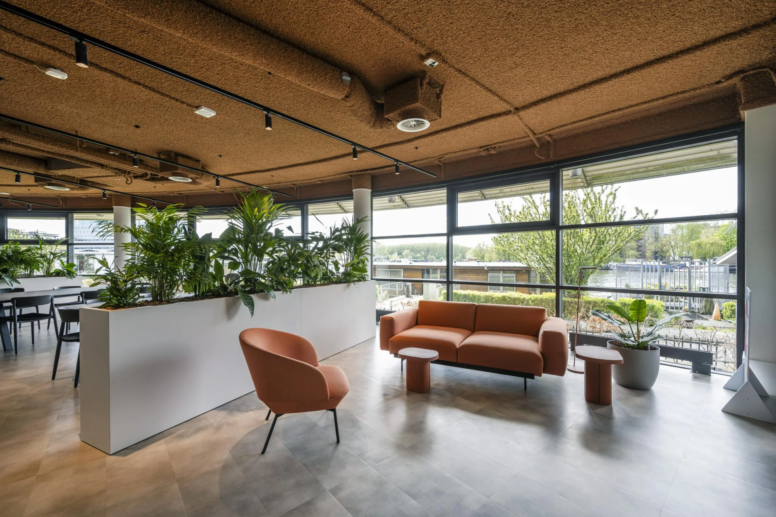 Modern lounge area at Omval with terracotta seating, indoor plants, and large windows overlooking a canal.
