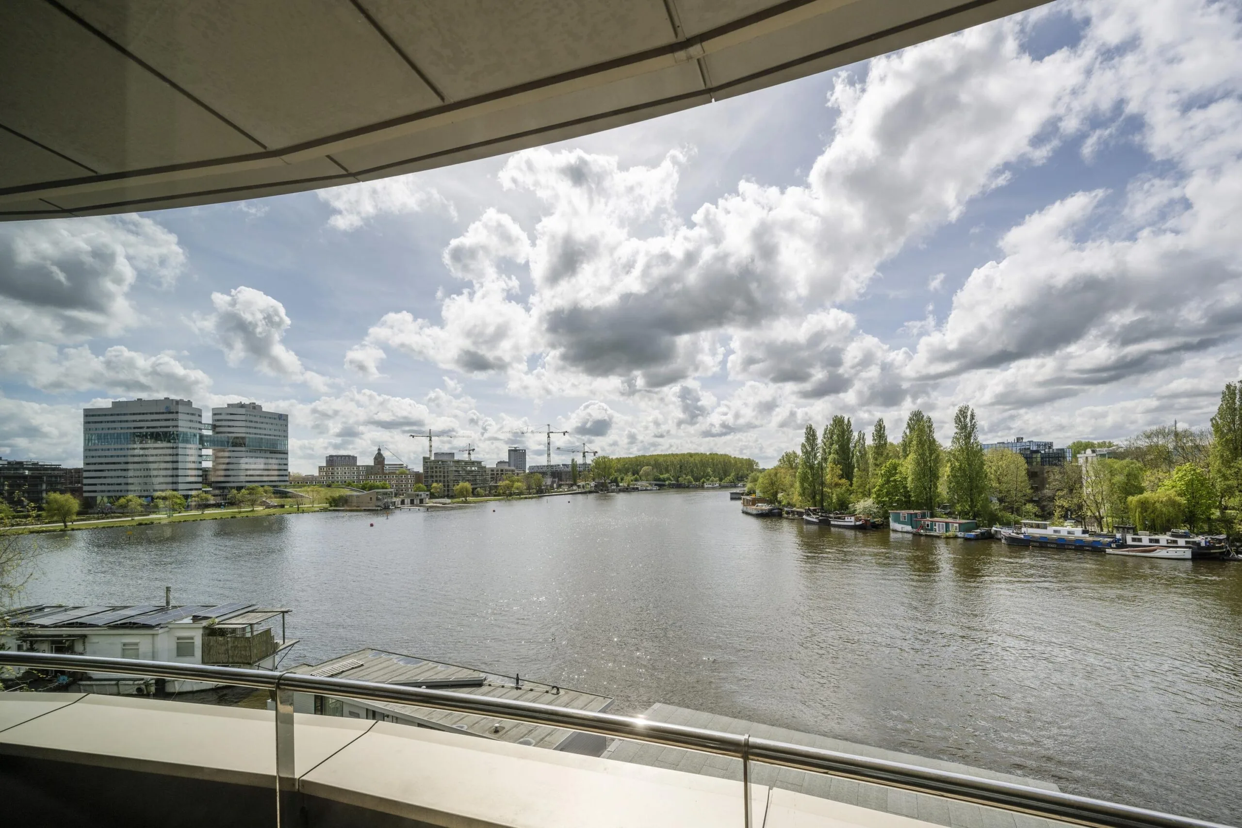 View of the Amstel River in the Omval district of Amsterdam with modern office buildings, houseboats, and a cloudy sky.