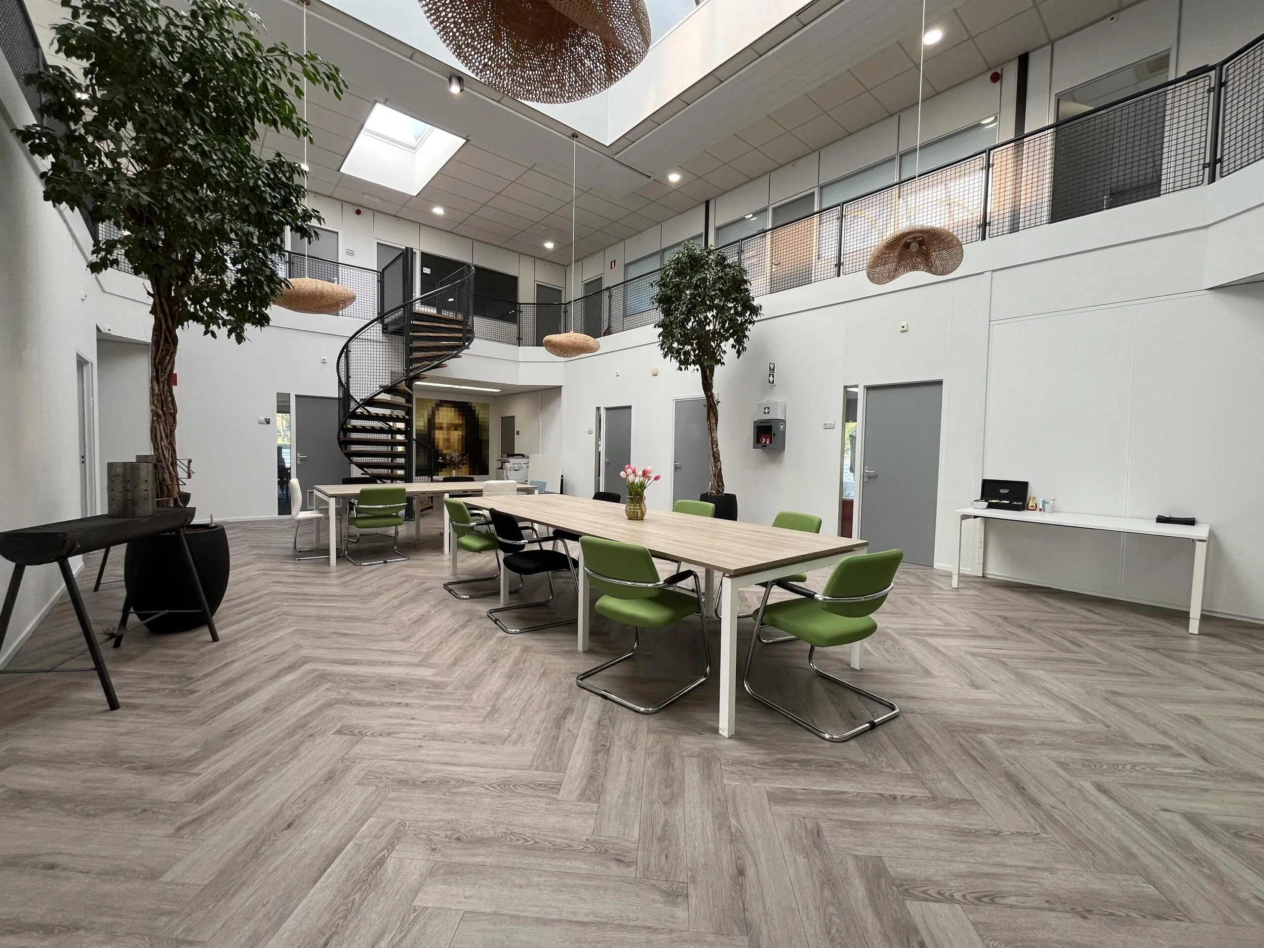 Modern open office space at Archimedesbaan with a spiral staircase, green chairs, and indoor trees under a skylight.
