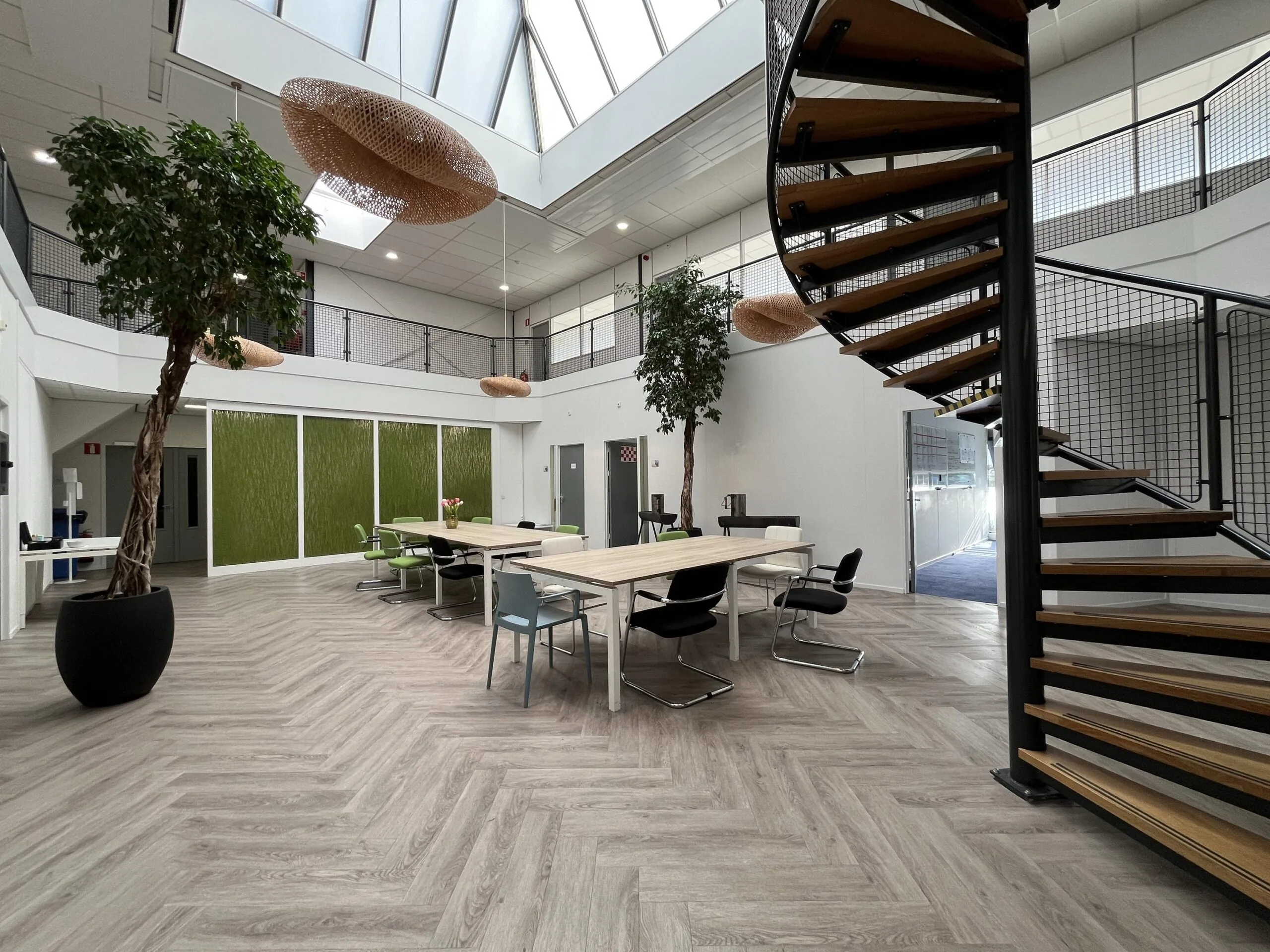 Modern open office space at Archimedesbaan with large skylight, spiral staircase, shared work tables, and indoor plants.