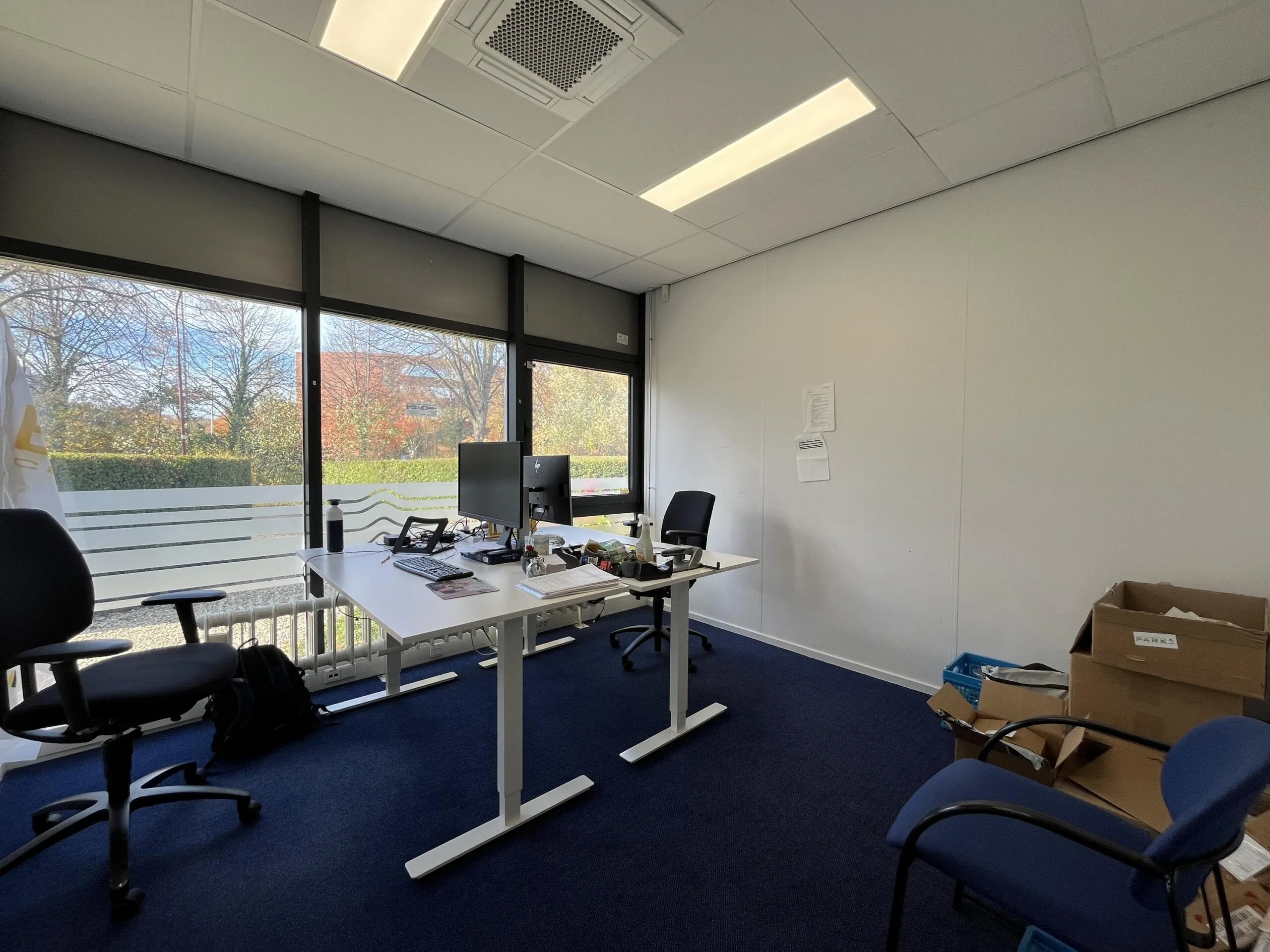 Modern office room on Archimedesbaan with two desks, office chairs, and large windows overlooking trees.