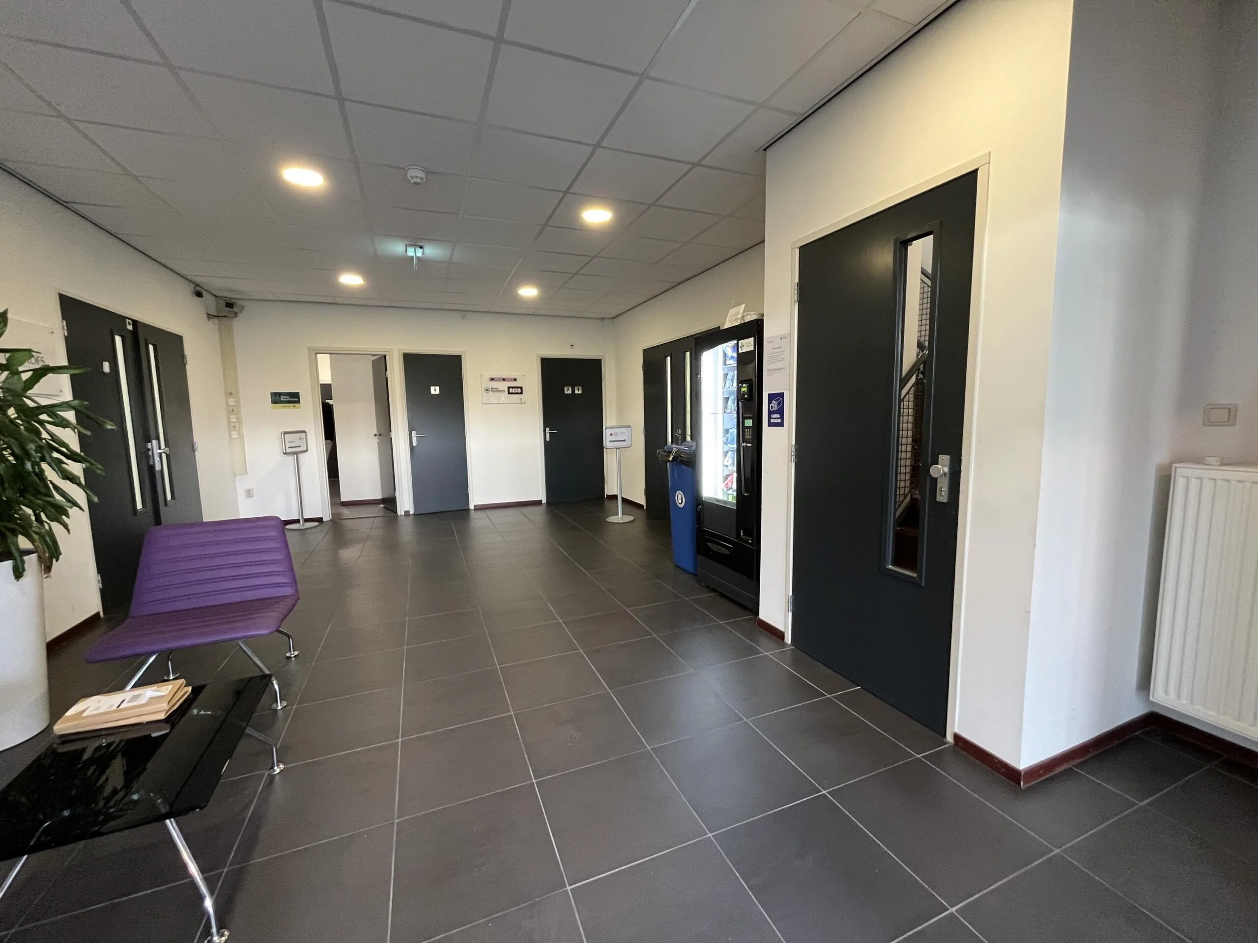 Modern hallway at Archimedesbaan featuring dark doors, a vending machine, a purple bench, and a black tiled floor.