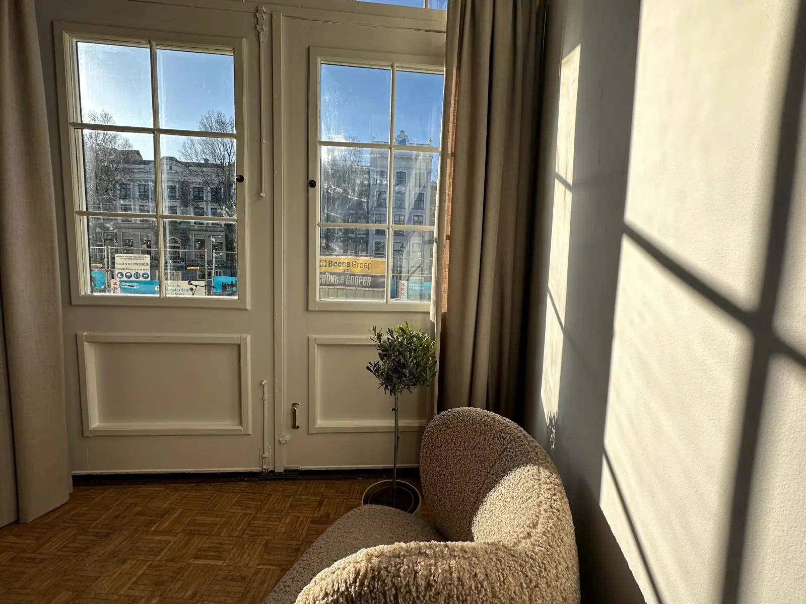 Sunlight streams through French doors overlooking Herengracht, casting shadows on the wall of a cozy interior with a curved armchair and a potted plant.