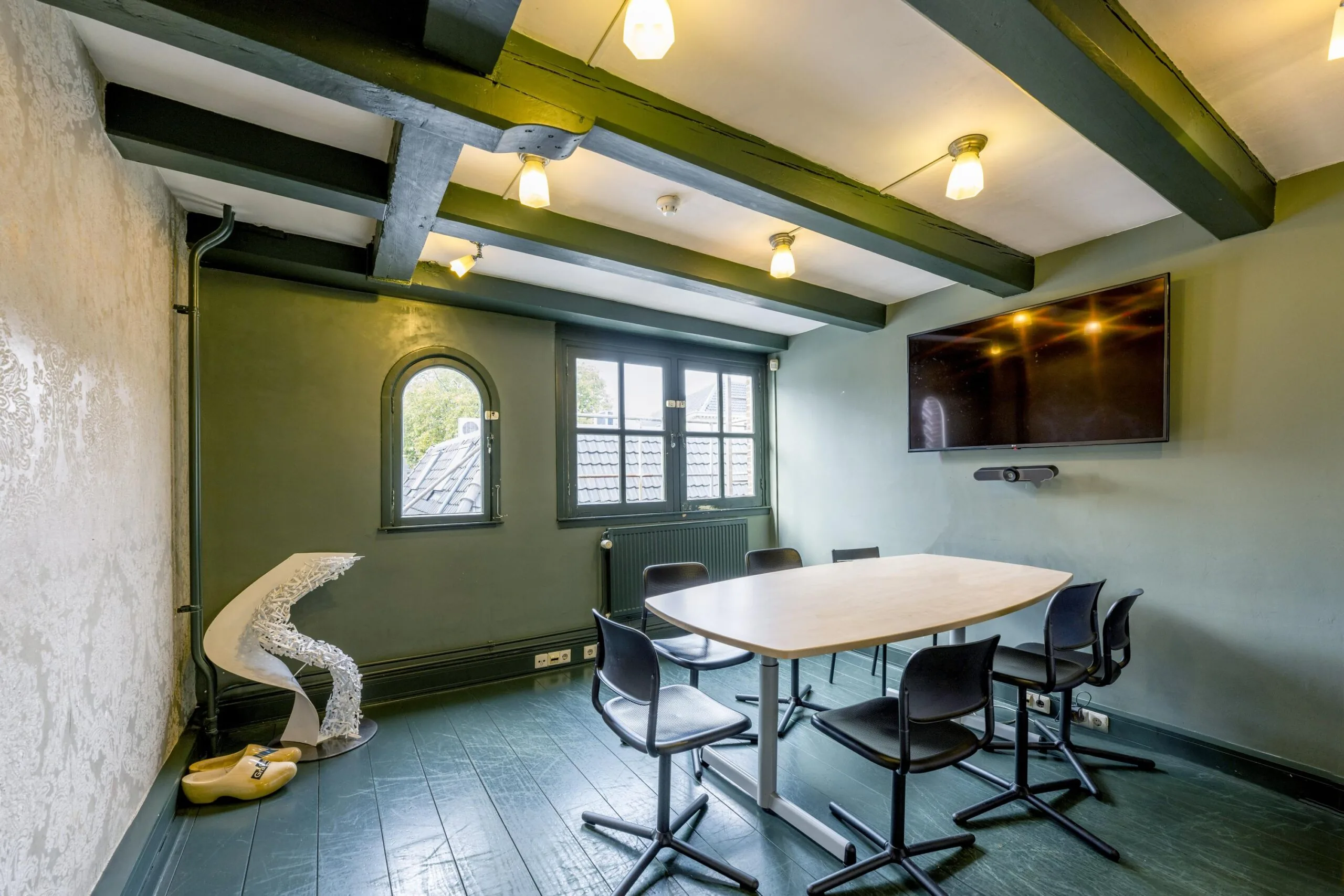 Modern meeting room on Leidsegracht with a wooden table, six black chairs, wall-mounted TV, and exposed ceiling beams.