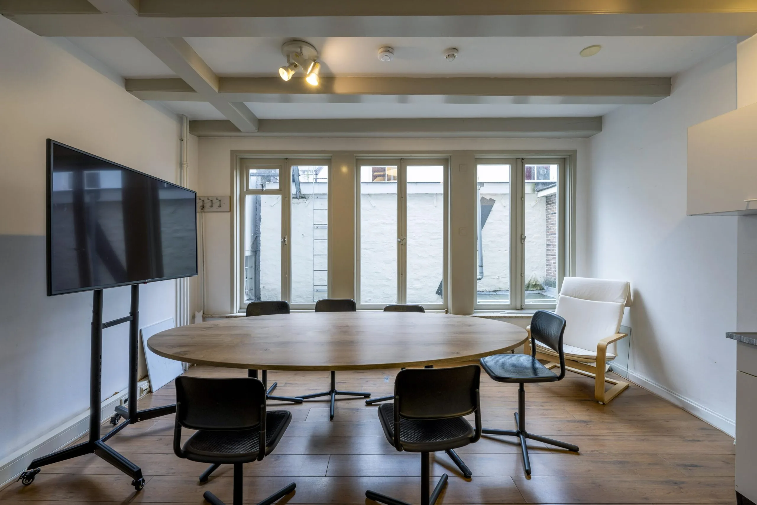 Modern meeting room with a round wooden table, black chairs, a large TV screen, and tall windows at Leidsegracht.