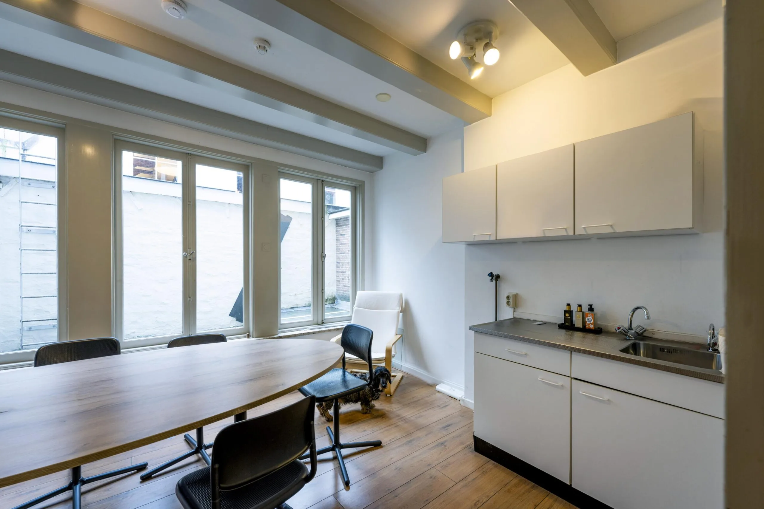 Bright kitchenette and meeting area on Leidsegracht with a wooden table, black chairs, and a small dog near the window.
