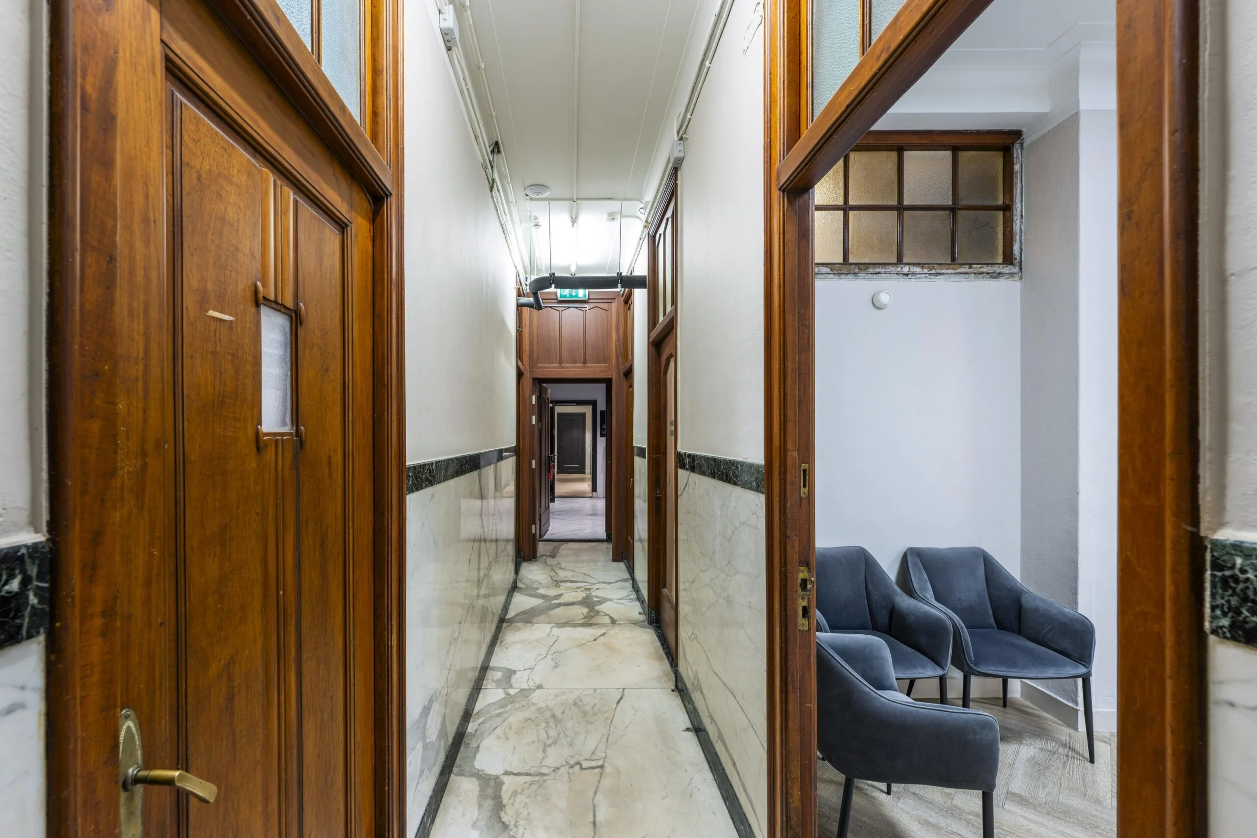 Narrow hallway in the Westermarkt building with marble walls, wooden doors, and a small waiting area with three blue chairs.