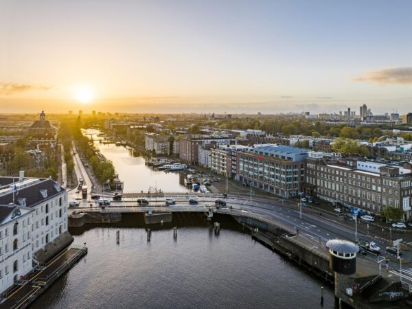 Aerial view of the Nieuwe Vaart canal in Amsterdam at sunrise, with surrounding buildings and a bridge in the foreground.