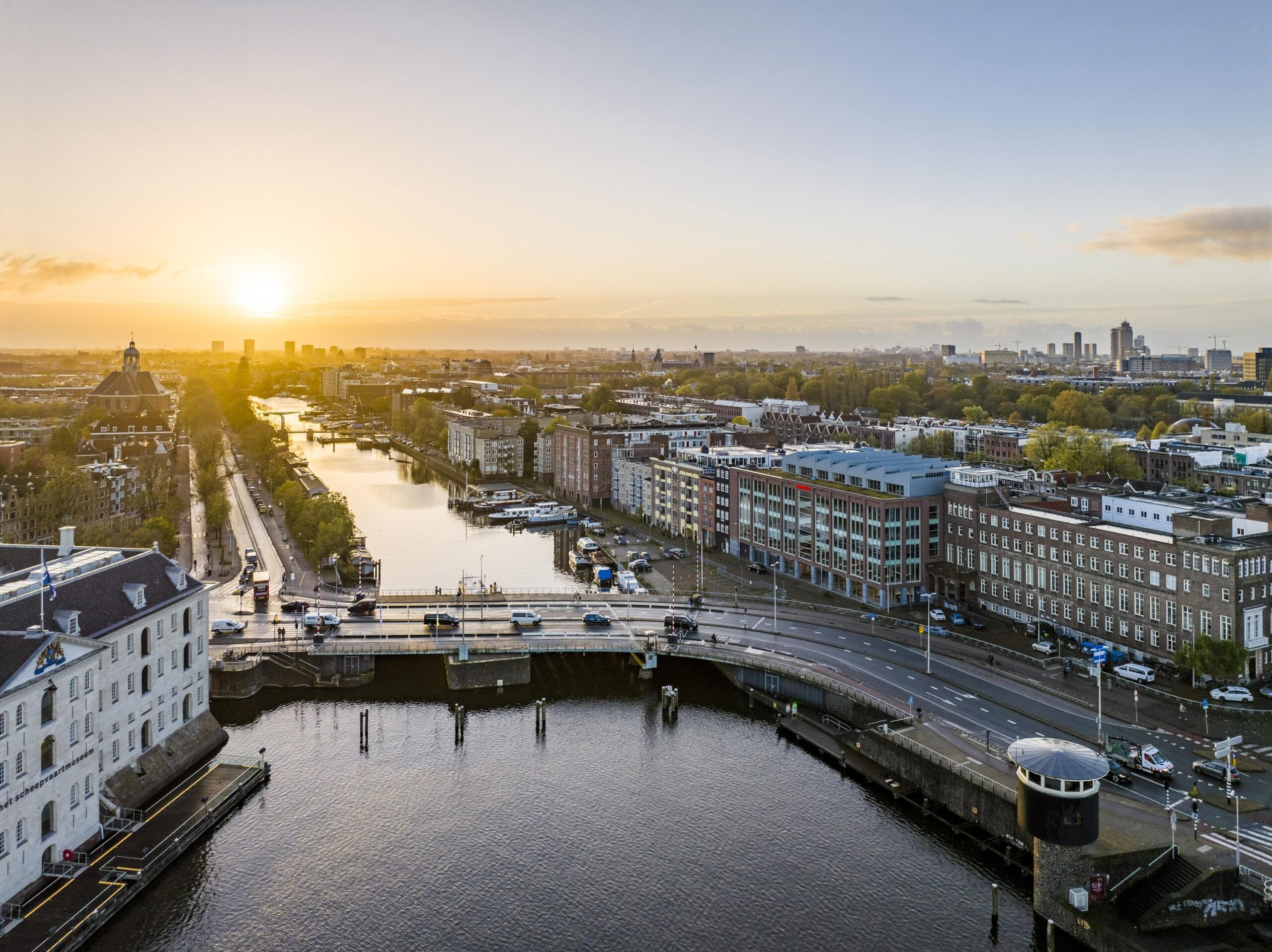Aerial view of the Nieuwe Vaart canal in Amsterdam at sunrise, with surrounding buildings and a bridge in the foreground.