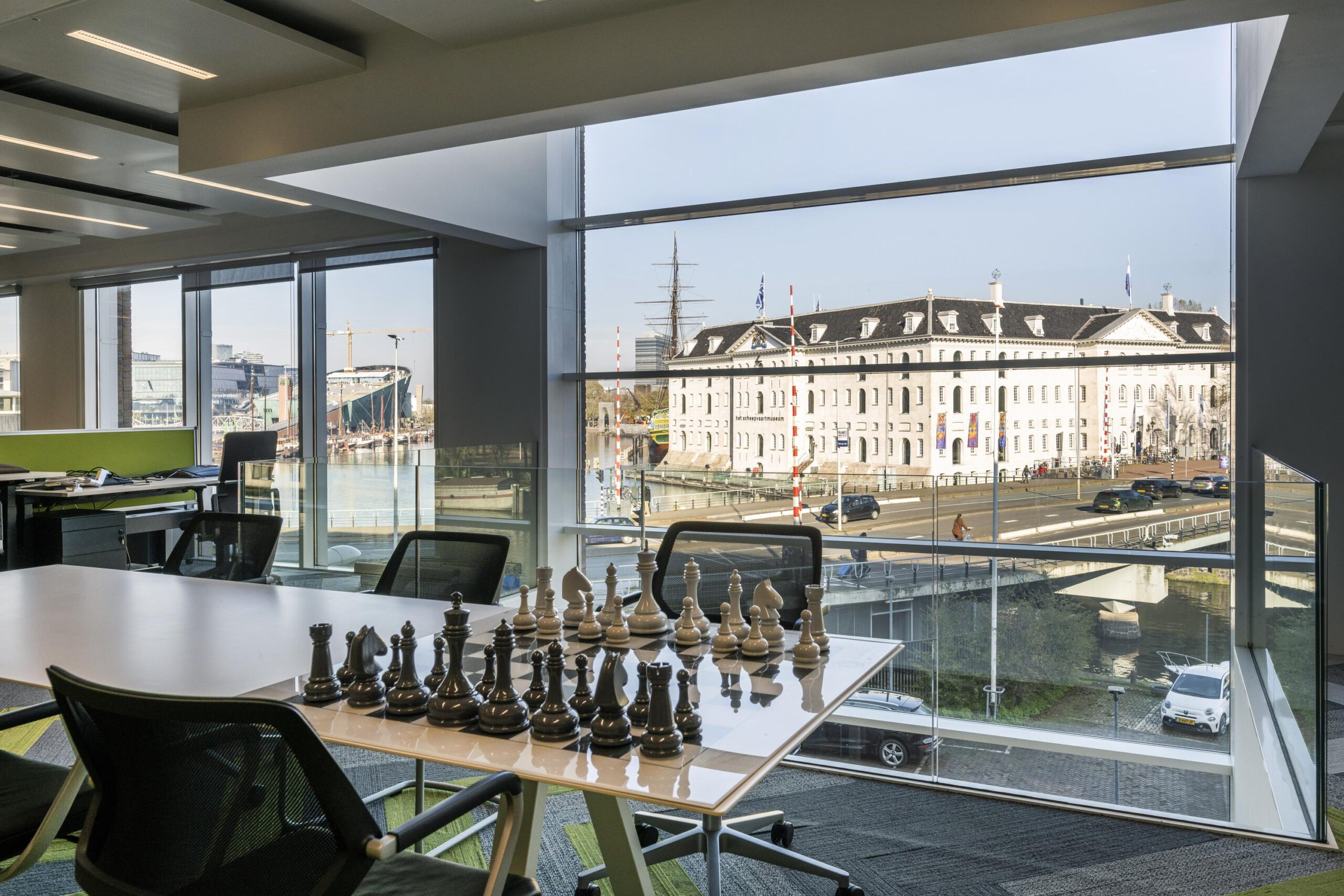 Modern office interior with a chessboard on the table, overlooking the Nieuwe Vaart canal and the Maritime Museum in Amsterdam through large glass windows.