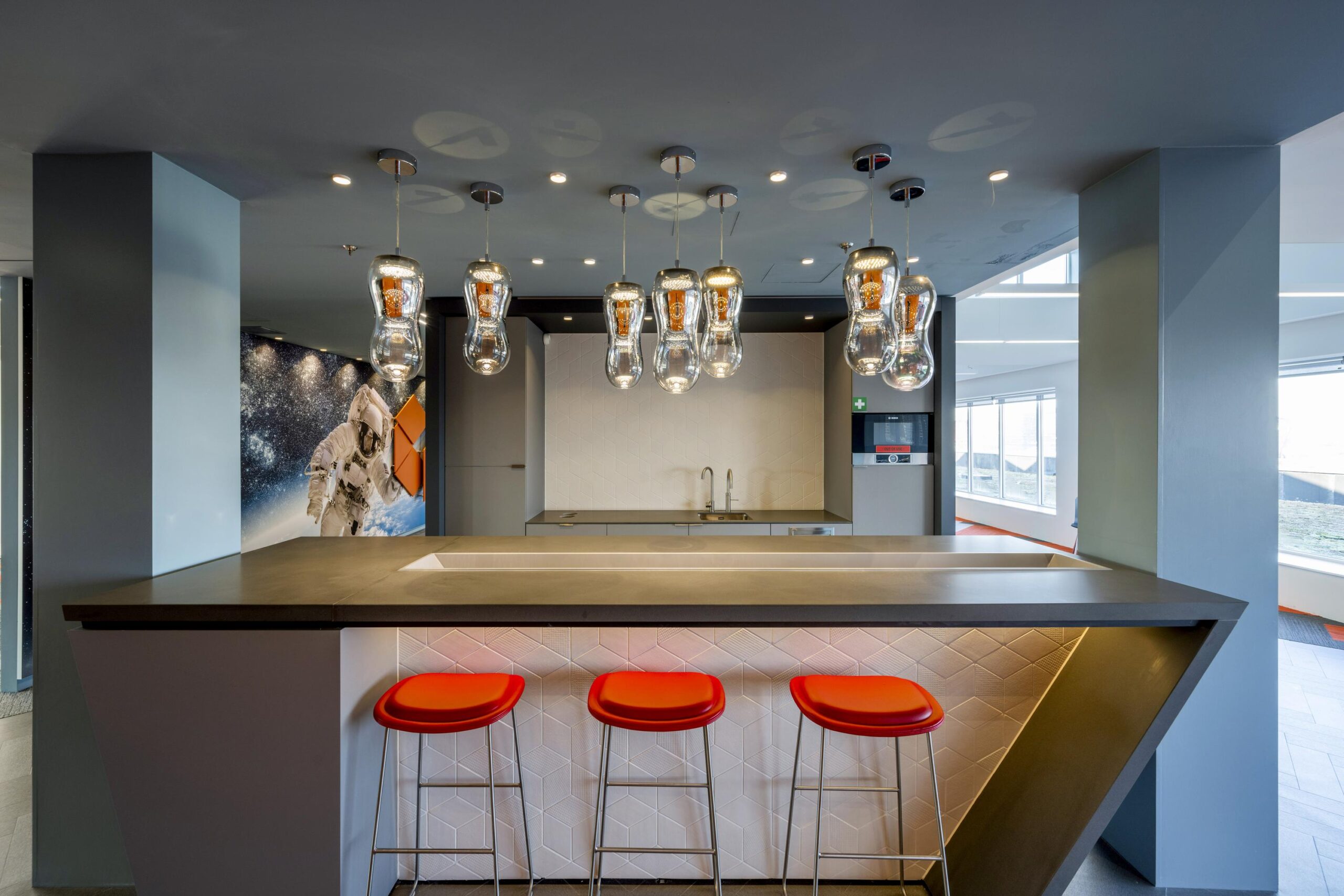 Modern kitchen area with a geometric island, red bar stools, hanging light fixtures, and a space-themed wall mural featuring an astronaut.
