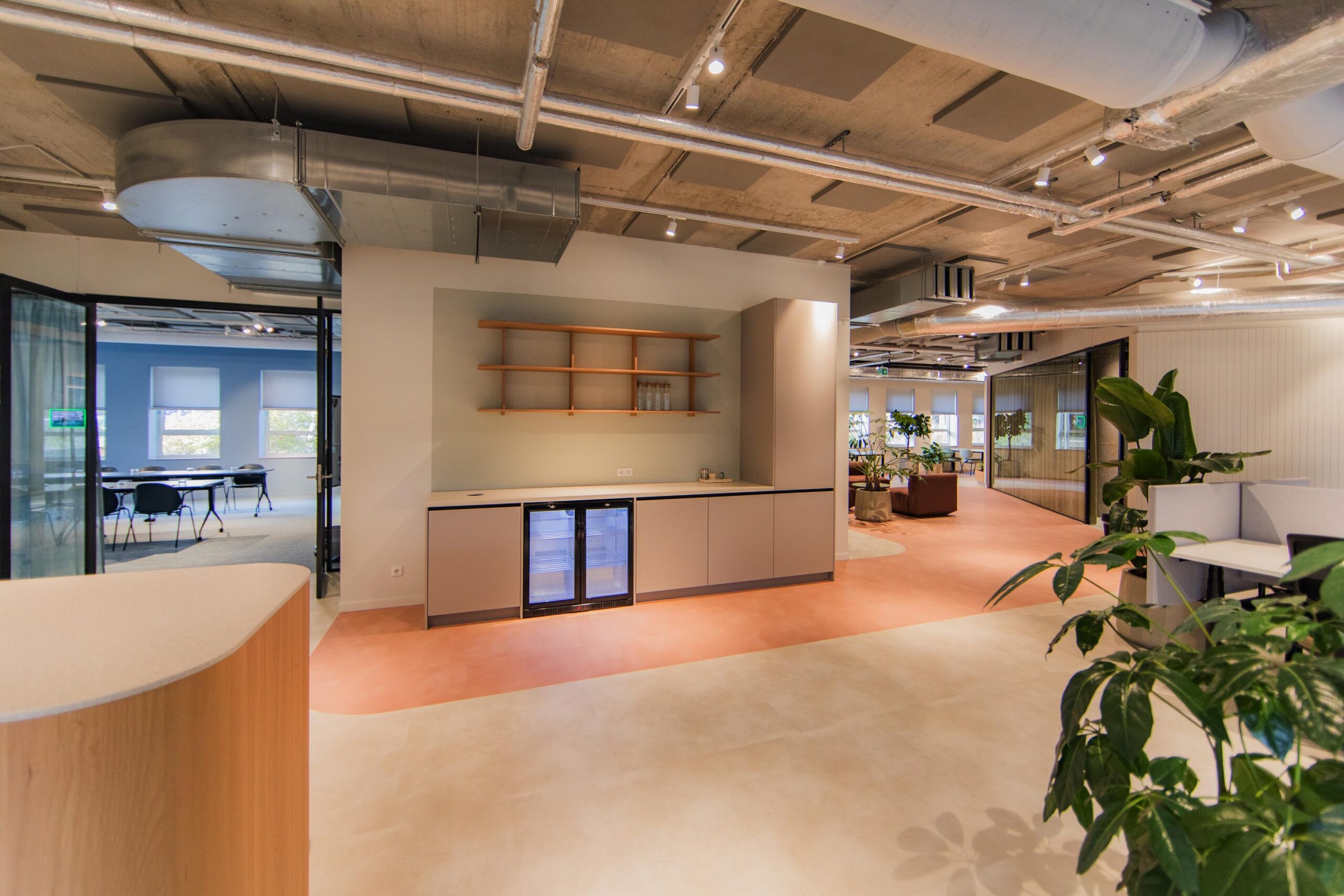 Modern office interior at Hullenbergweg featuring an open kitchenette, meeting room, and lounge area with plants and industrial ceiling elements.
