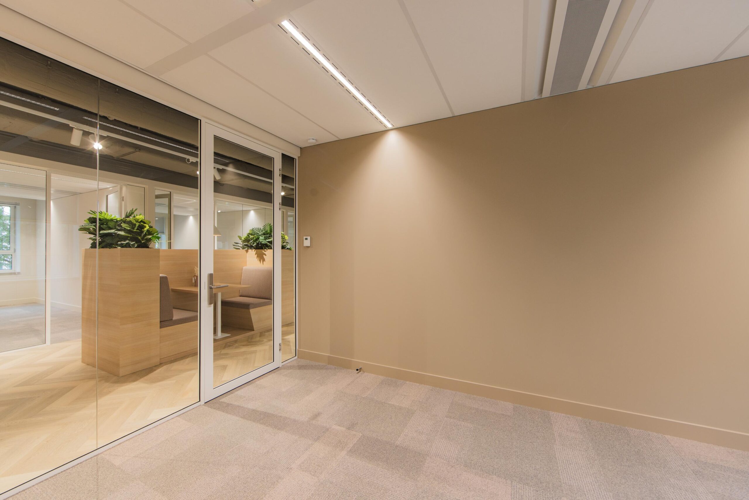 Modern office interior at Hullenbergweg with glass walls, beige carpeting, and wooden seating areas with built-in planters.