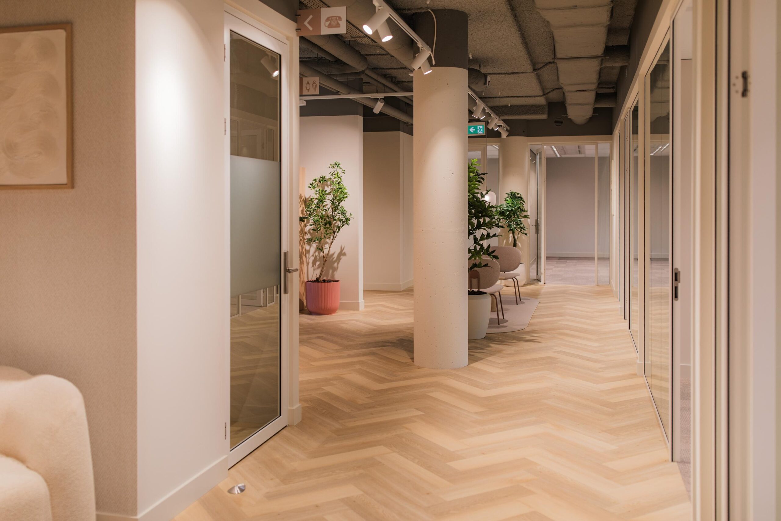 Modern office hallway at Hullenbergweg with wooden herringbone flooring, glass doors, and indoor plants.