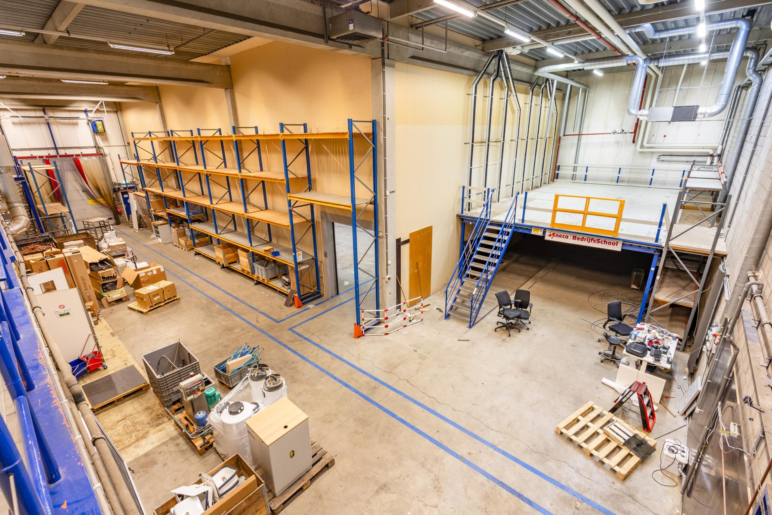 Een overzicht van een opslagruimte met houten stellingen, een werkruimte op een verhoging en diverse materialen op de vloer.