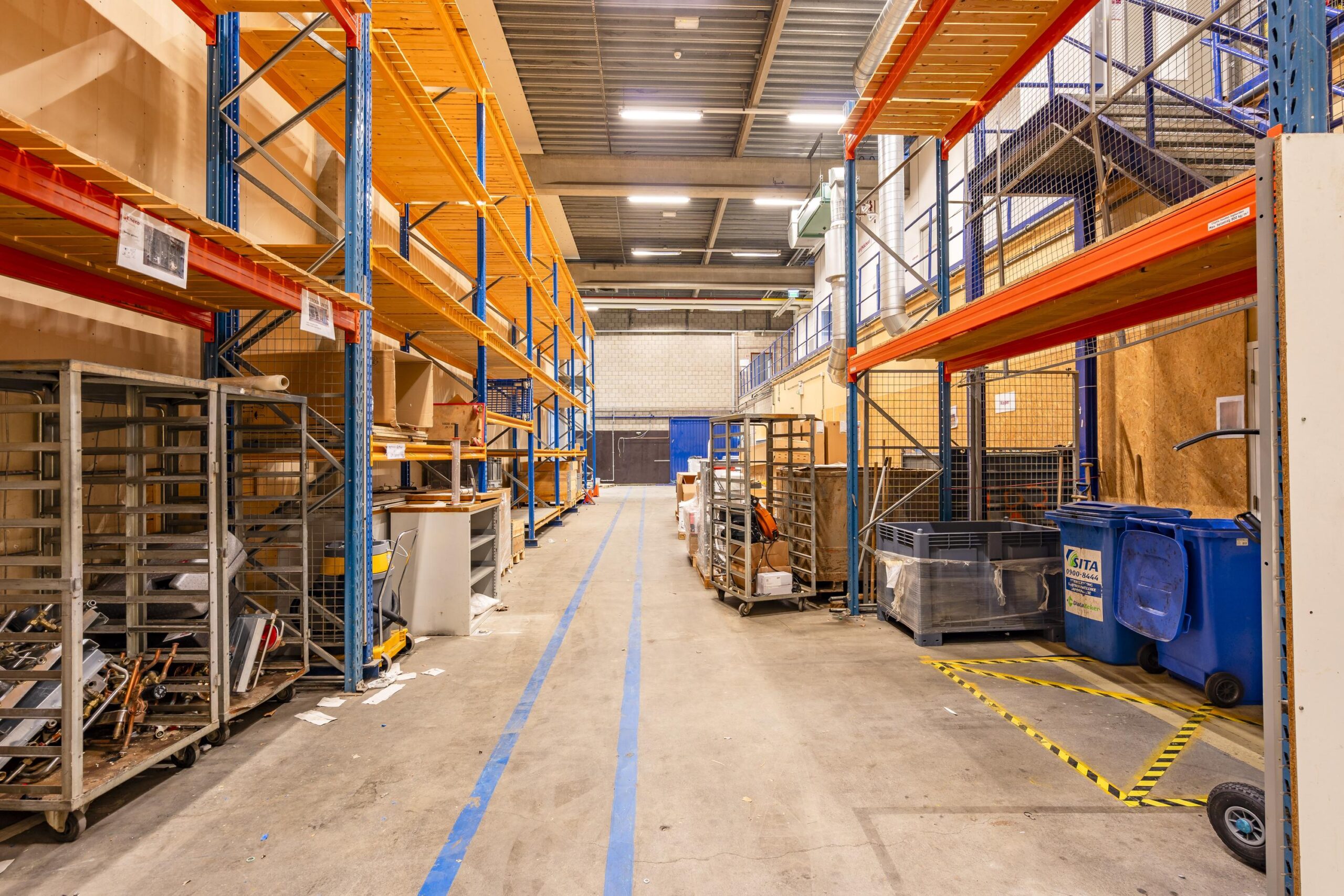 Een ruime gang in een opslagloods met metalen stellingen en diverse opgeslagen goederen.