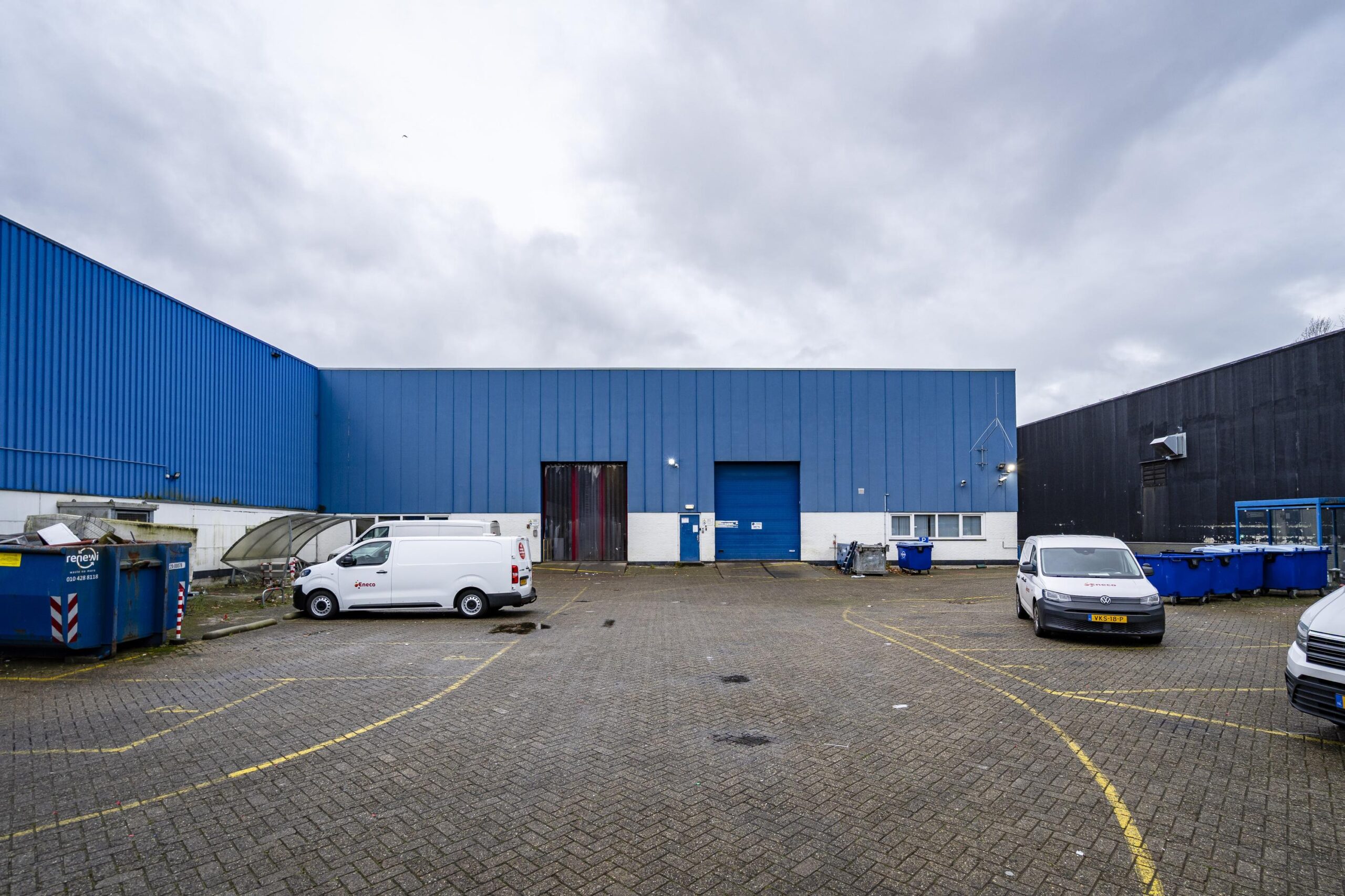 Blauwe industriële loods aan de s-Gravelandseweg met geparkeerde bedrijfswagens en containers op de voorgrond.