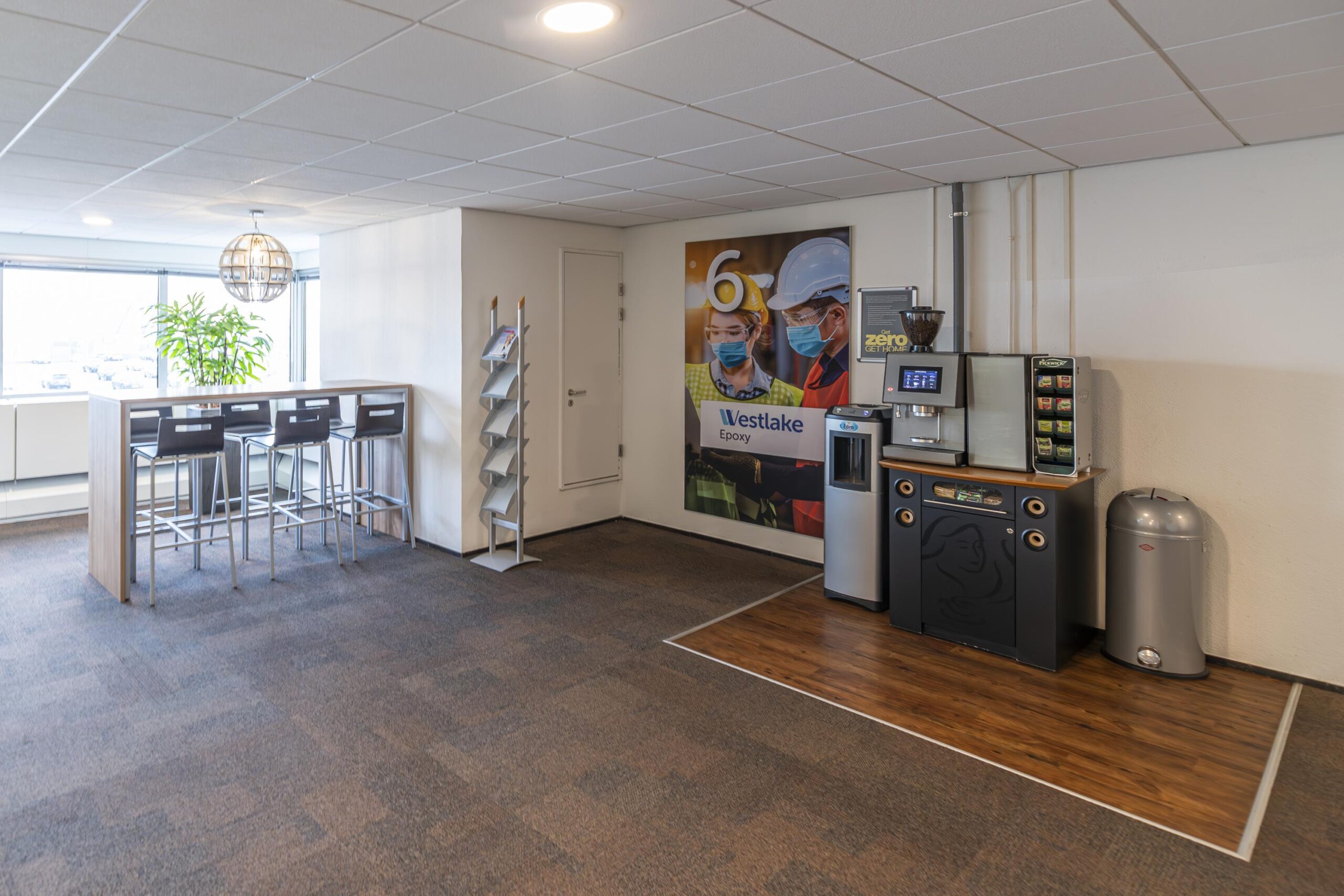 Kantinehoek met koffiemachine, hoge tafel met barkrukken en een poster van Westlake Epoxy in een bedrijfsruimte aan de Seattleweg.