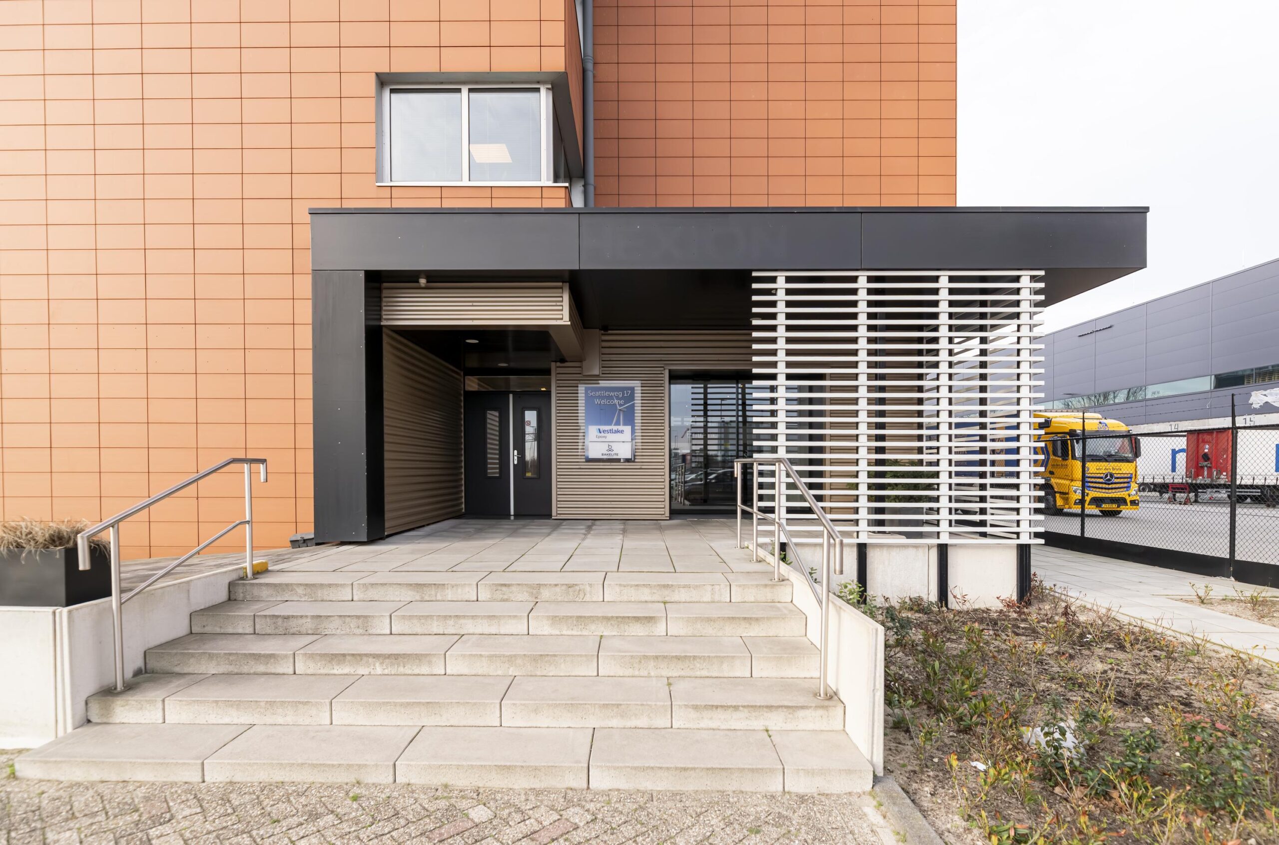 Ingang van een modern bedrijfsgebouw aan de Seattleweg met brede traptreden en een gele vrachtwagen op de achtergrond.