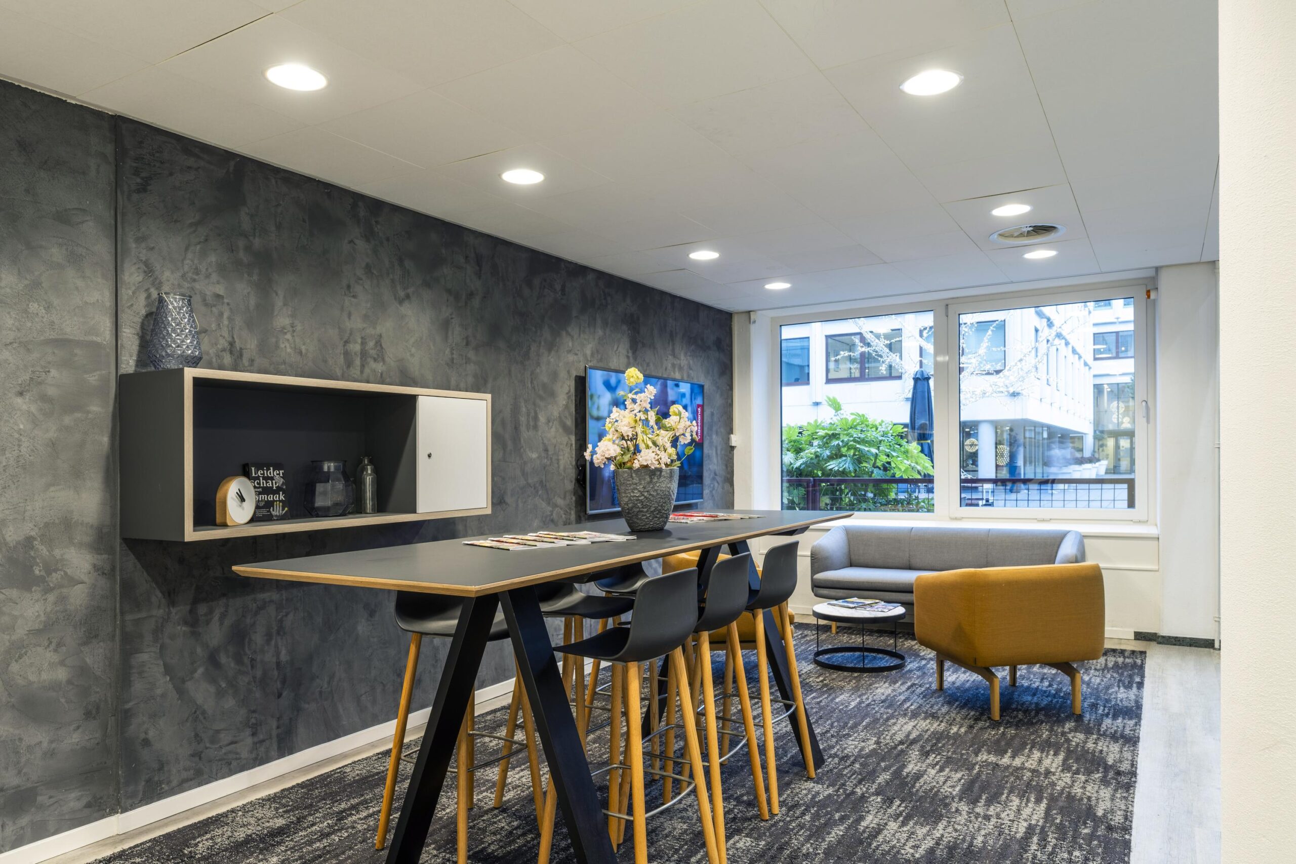 Moderne kantoorlounge met hoge tafel, barkrukken, zithoek en grote ramen met uitzicht op een binnentuin.