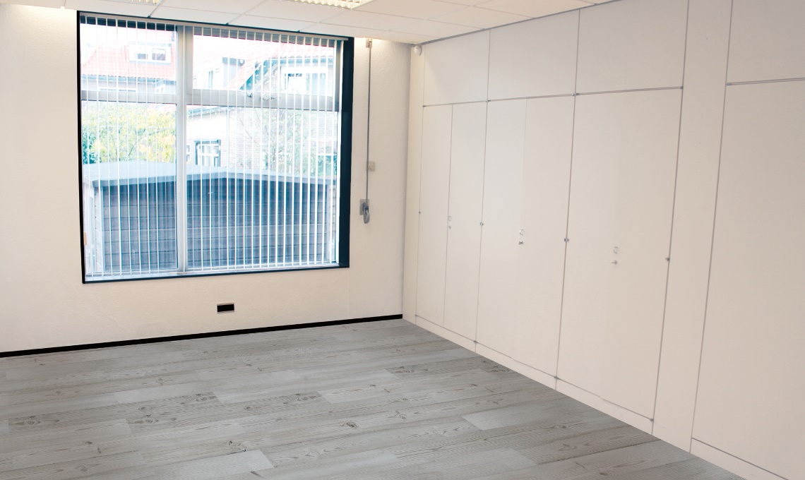 Lege, lichte kantoorruimte met groot raam, verticale jaloezieën en een wand met ingebouwde kasten aan Oosteinde.