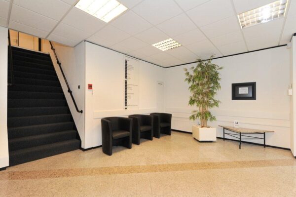 Wachtruimte in een bedrijfsgebouw met zwarte stoelen, een trap, een grote plant en een informatiebord aan de muur.