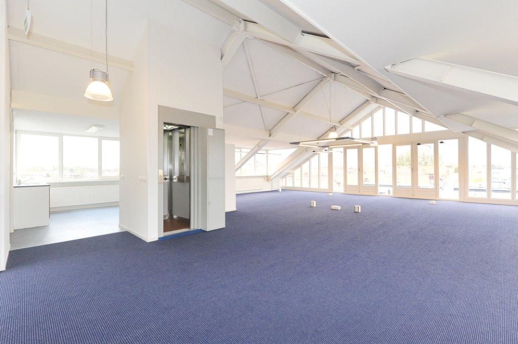 Ruime, lichte kantoorruimte met schuine plafonds, blauwe vloerbedekking, een lift en grote ramen aan Oosteinde.