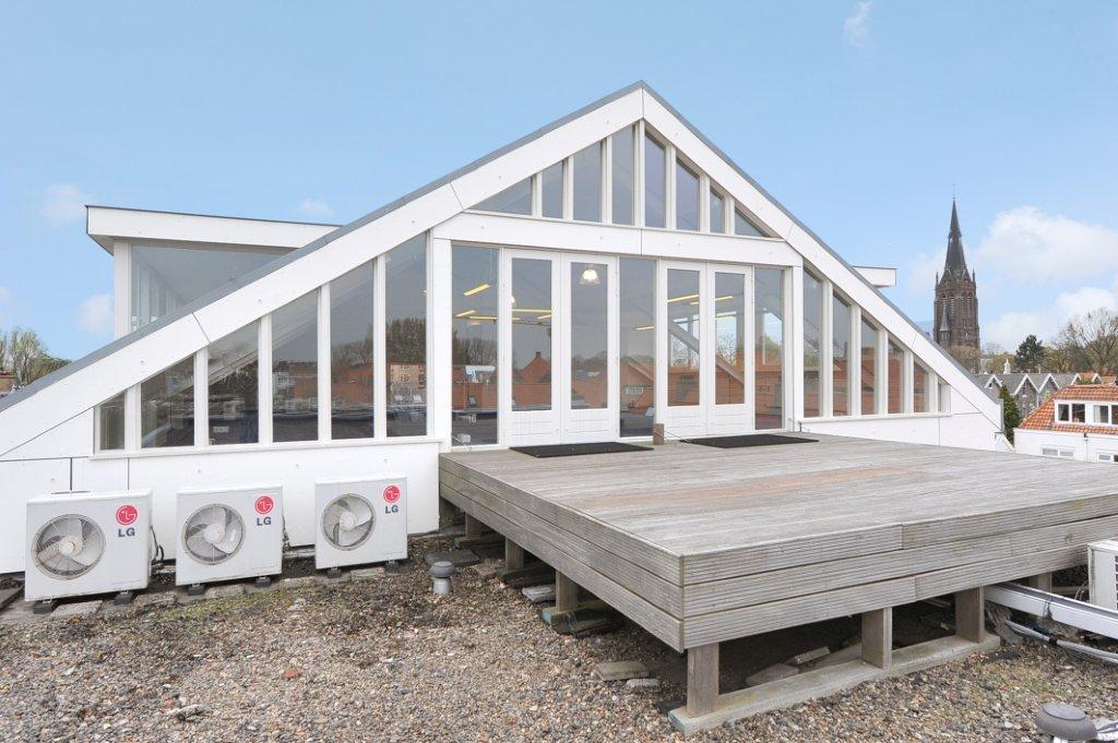 Modern wit gebouw met glazen gevel en houten dakterras op het Oosteinde, met zicht op een kerktoren op de achtergrond.