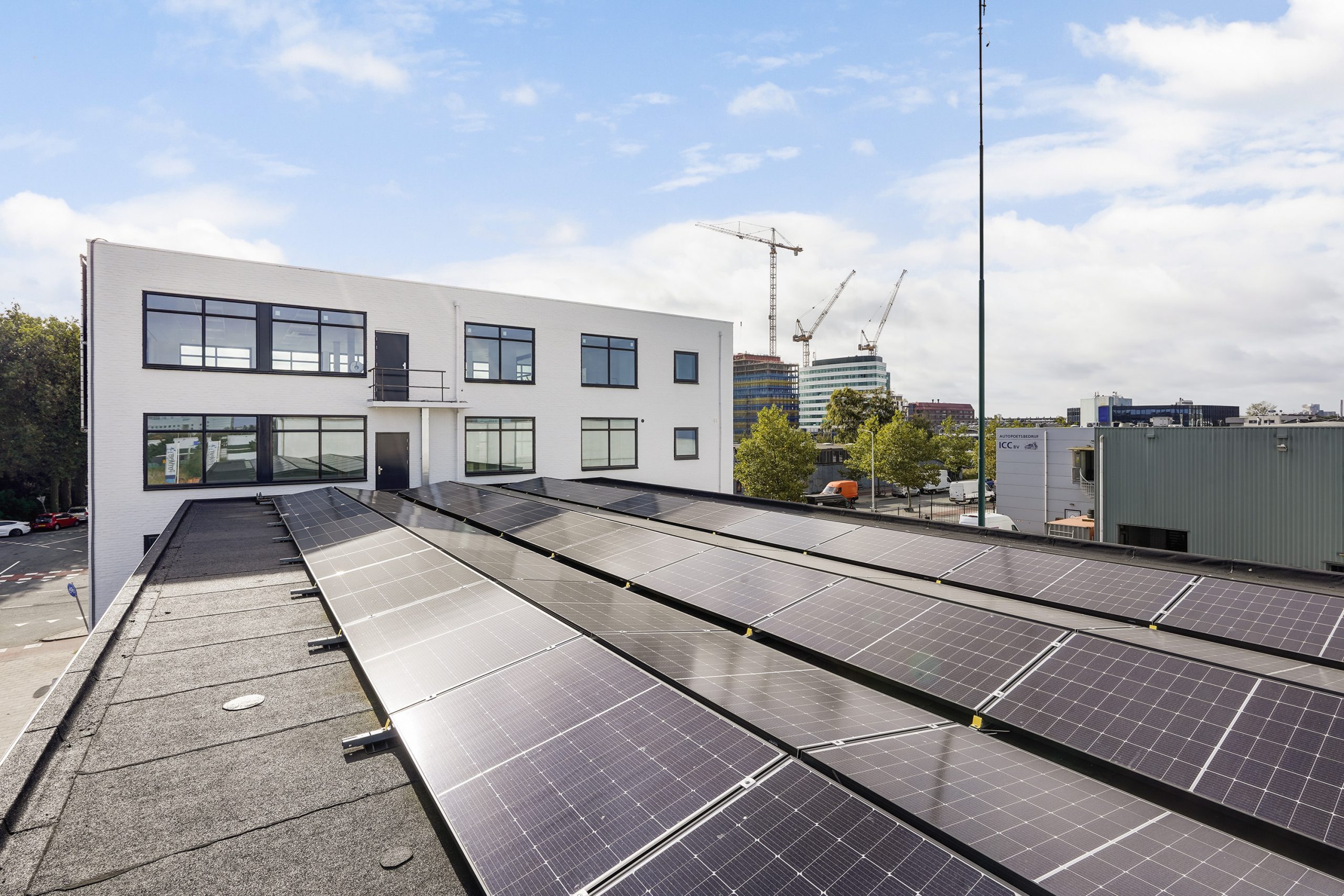 Plat dak van een wit kantoorgebouw aan de Mercuriusweg met zonnepanelen en uitzicht op een stedelijke omgeving met hijskranen.