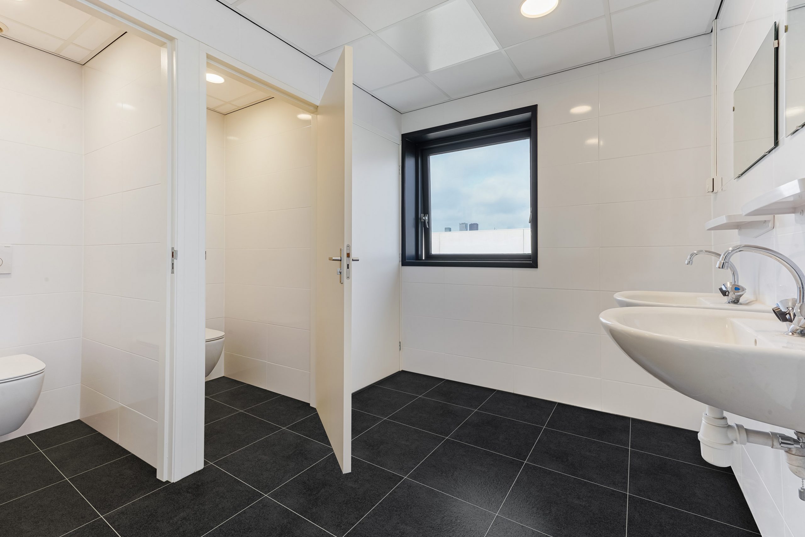 Moderne, wit betegelde toiletruimte met twee afgesloten toiletcabines, twee wastafels en een raam met zwart kozijn.