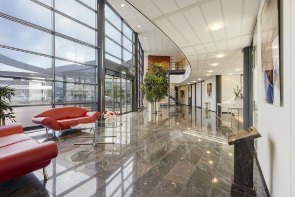 Moderne entreehal van een kantoorgebouw aan de Laan van ’s-Gravenmade met marmeren vloer, glazen gevel en rode designmeubels.